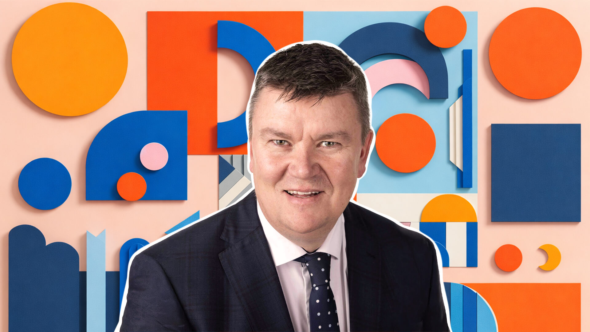 Person in a dark suit and tie seated against a colorful abstract background of geometric shapes.