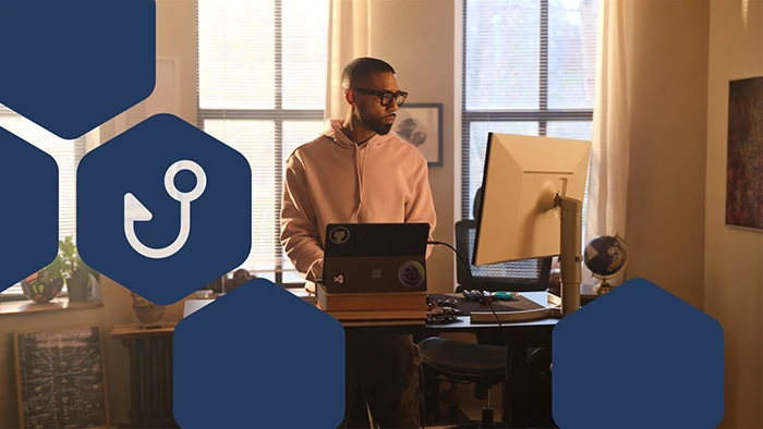 Person working at a standing desk with a laptop and monitor in a sunlit room.