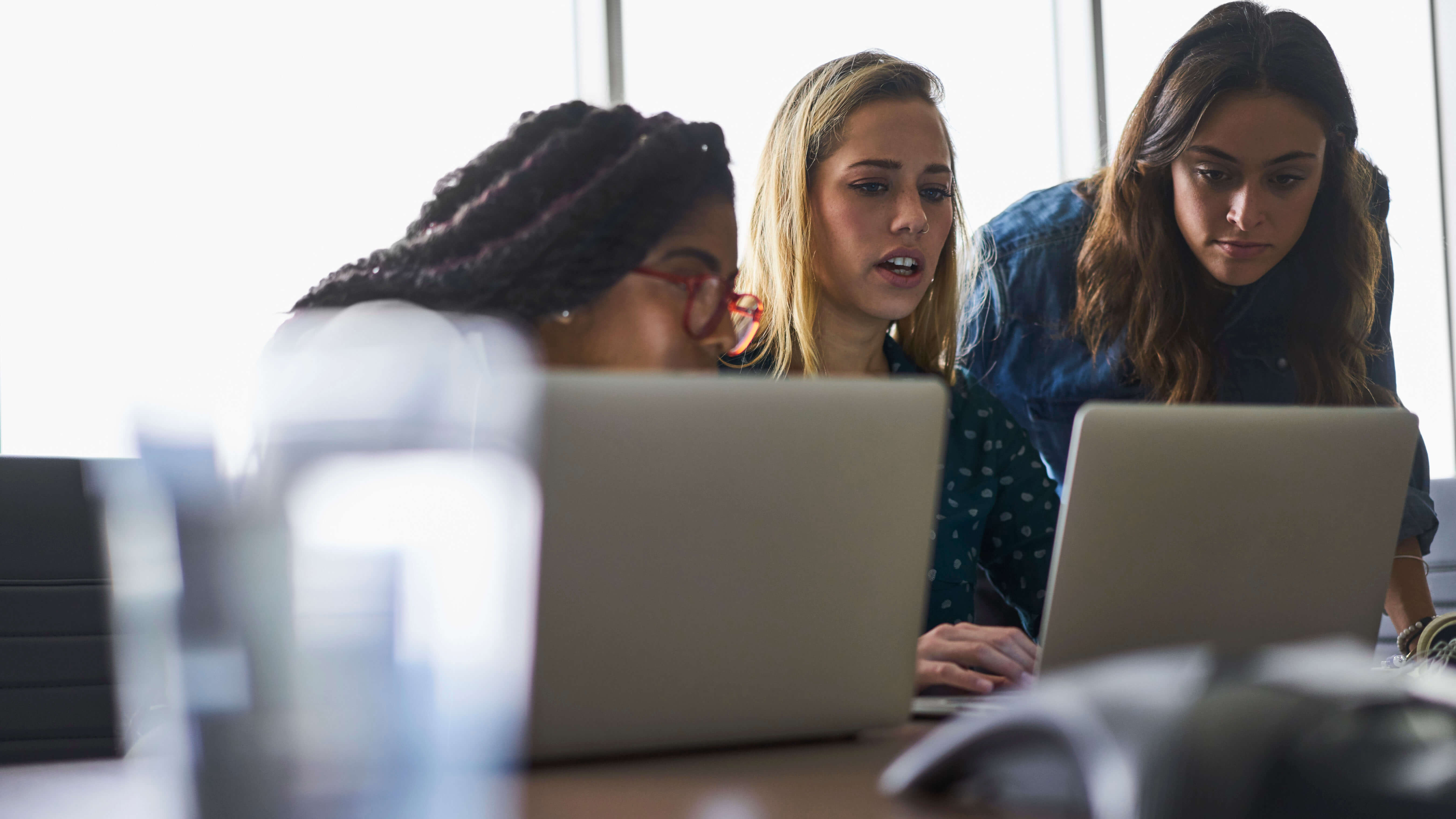 Three people collaborating at a table, looking at laptops in a bright office setting.