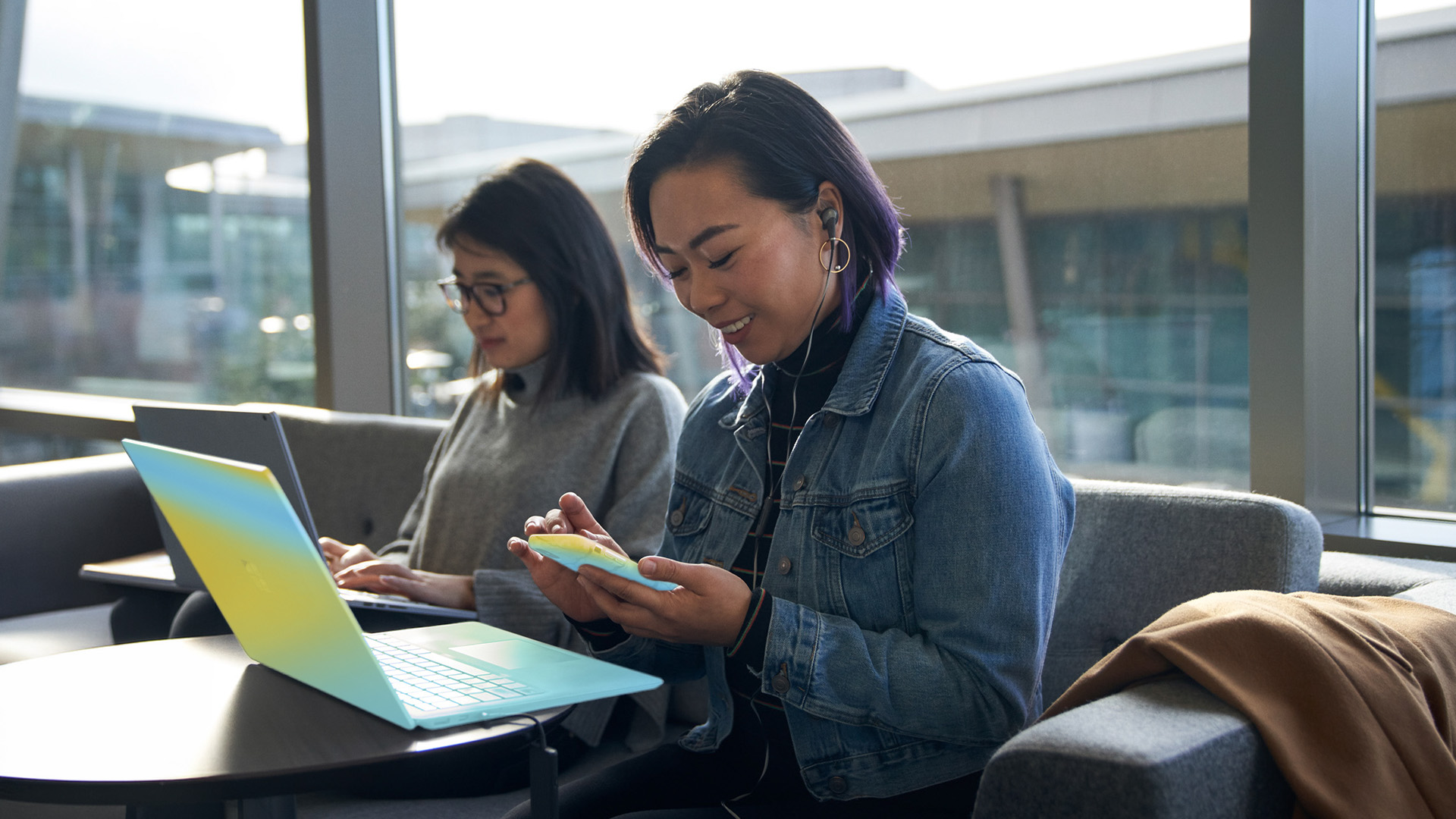 Two people seated on a couch, one working on a laptop and the other using a smartphone in a bright indoor lounge.