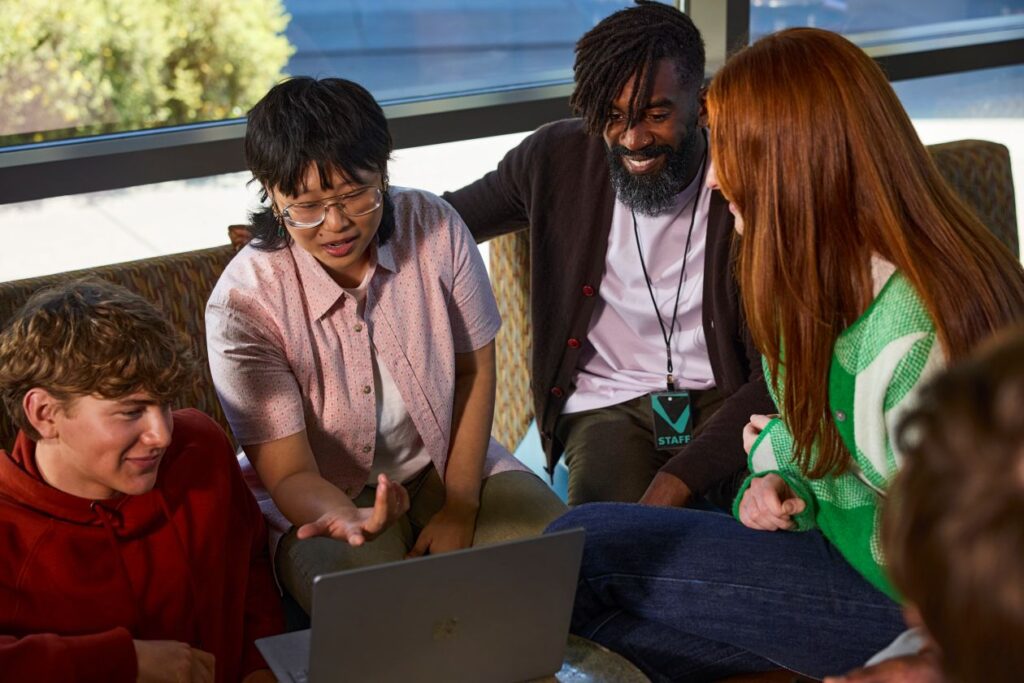 Group of people sitting together, looking at a laptop and discussing something on screen.