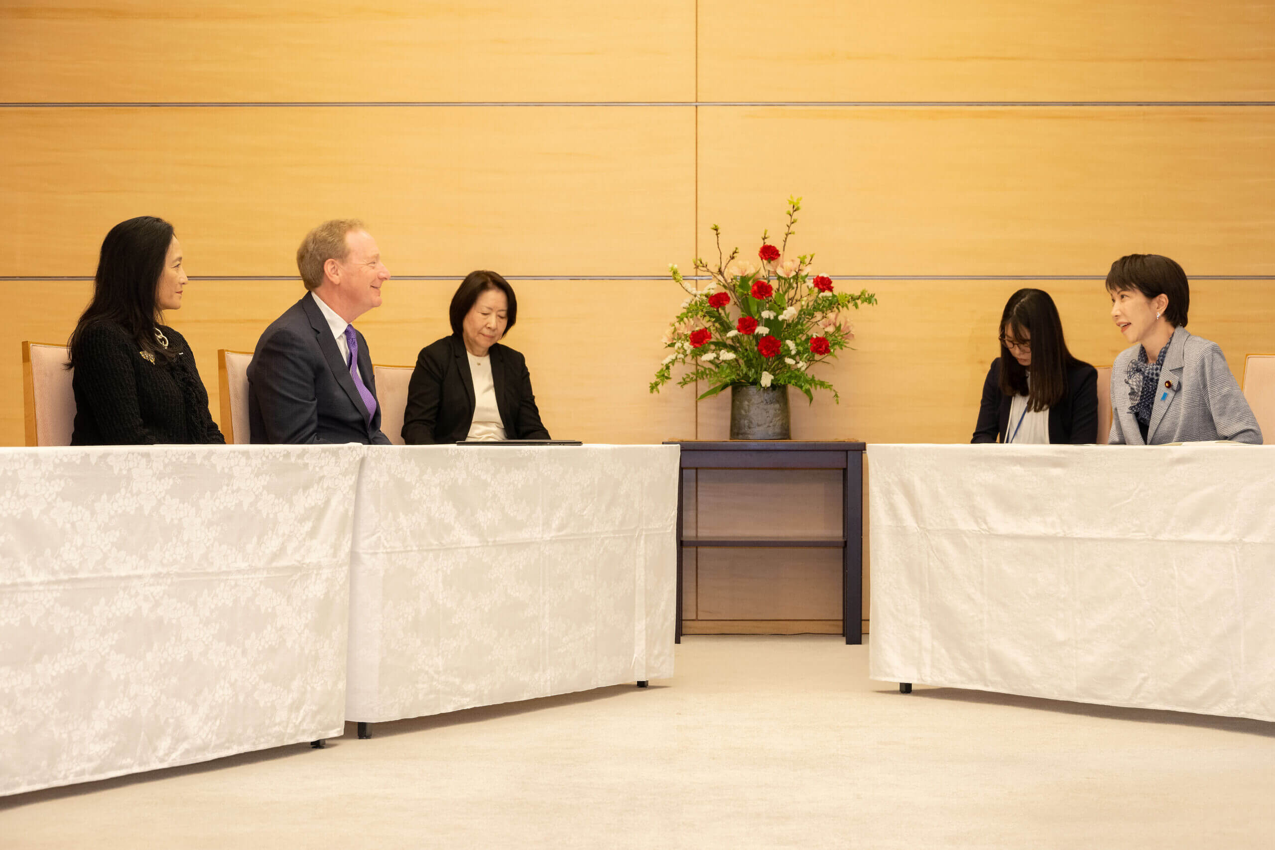 Six people seated across two tables in a formal meeting, with a flower arrangement between them.