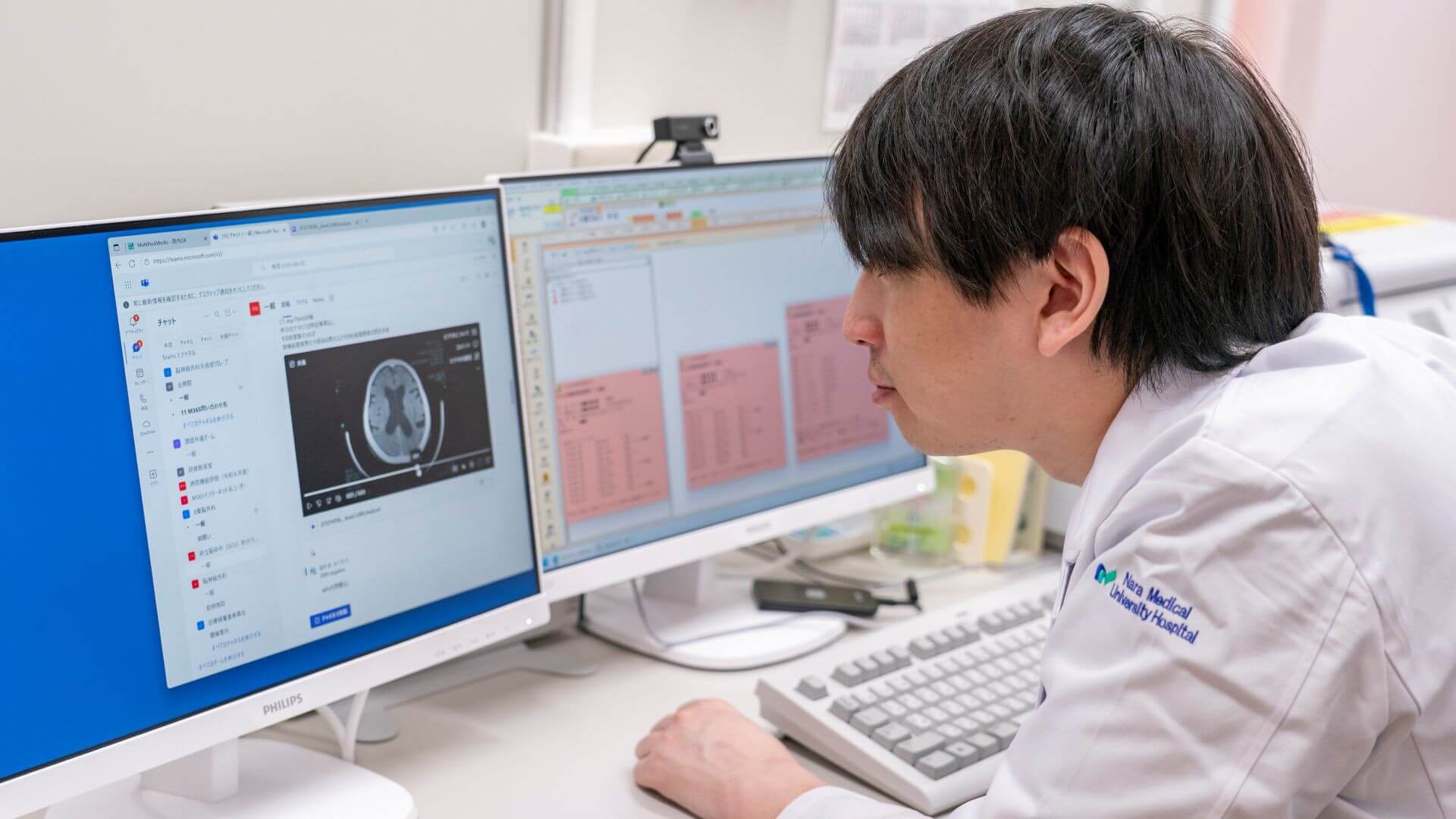 Person in a lab coat reviews medical imaging scans on a dual‑monitor computer workstation.