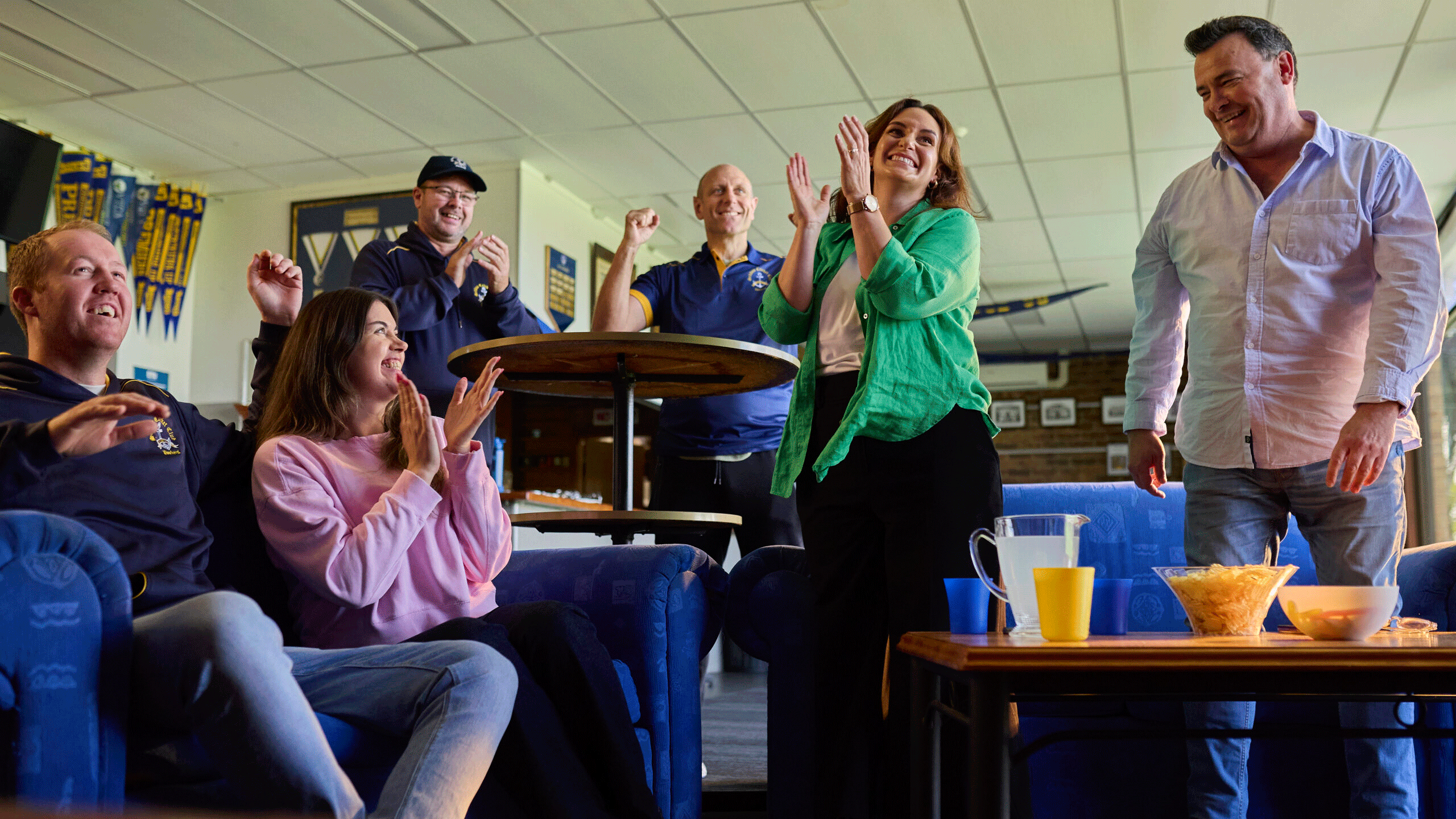 Group of six people in a casual lounge cheering and clapping around a table with drinks and snacks.