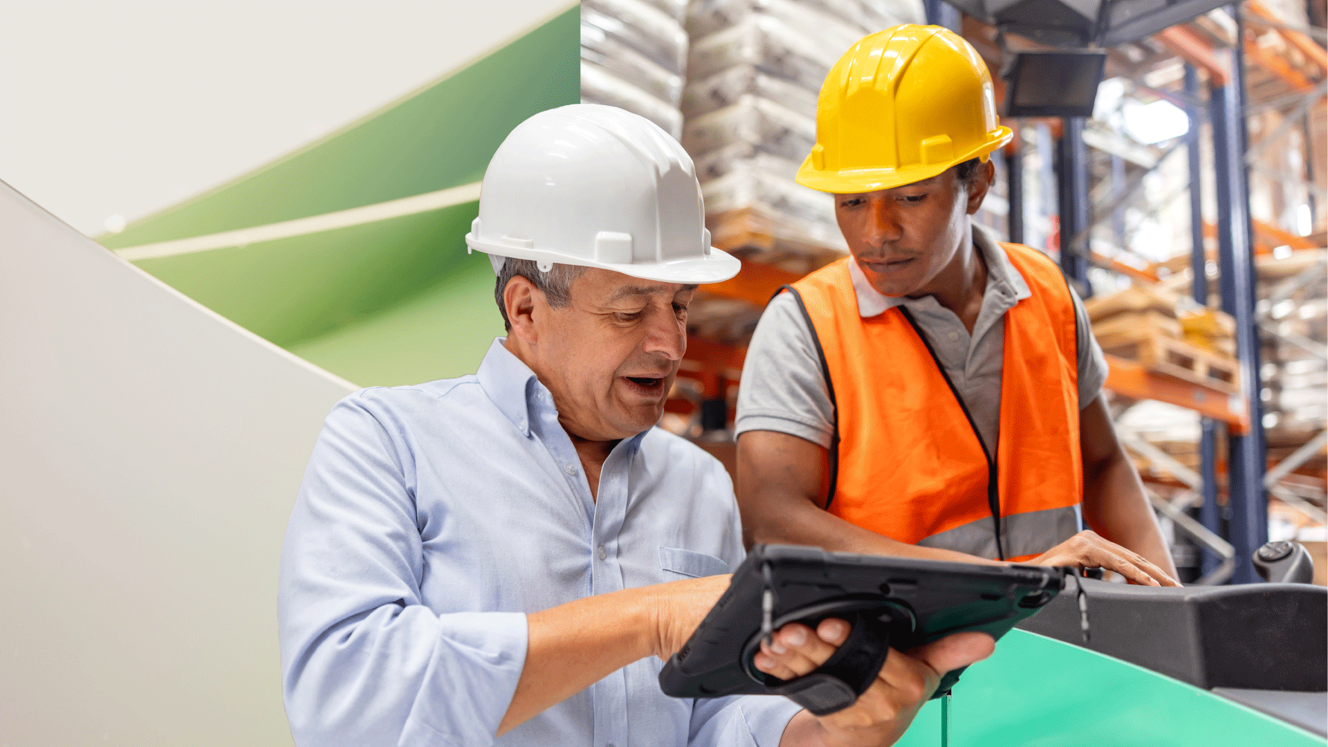 Two workers wearing hard hats review information on a tablet in a warehouse.