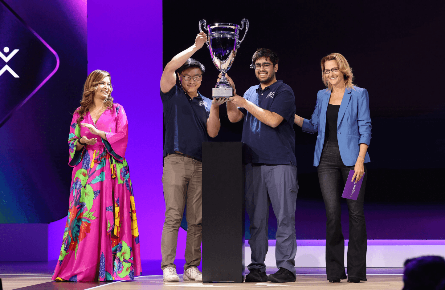 Two people on stage lift a large trophy as two others stand beside them; purple-lit backdrop and podium in center.