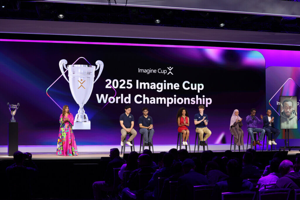 Wide stage with “2025 Imagine Cup World Championship” text; presenter stands beside seated finalists as audience looks on.