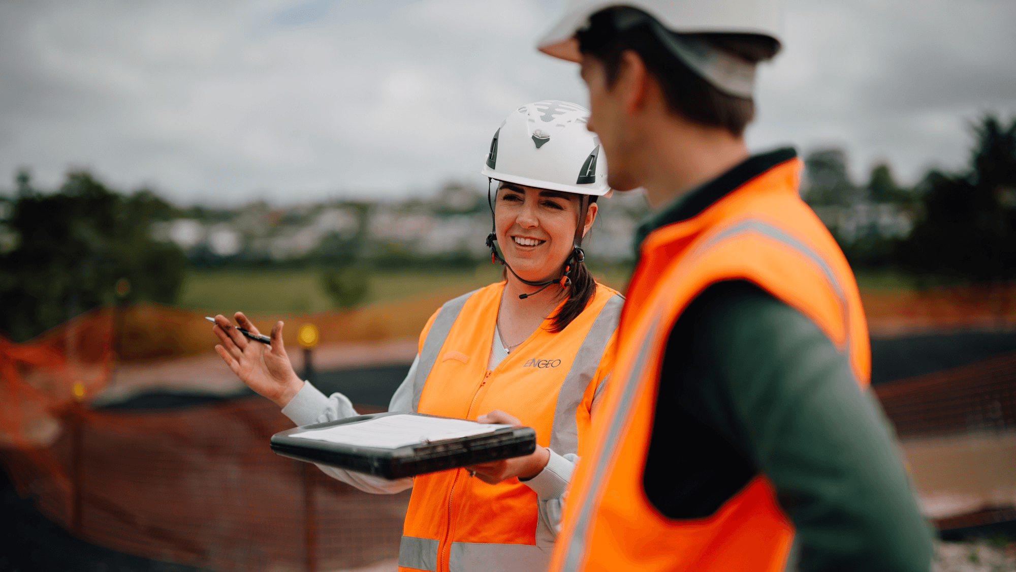 Person in a hard hat and safety vest discussing work with a colleague at a construction site.