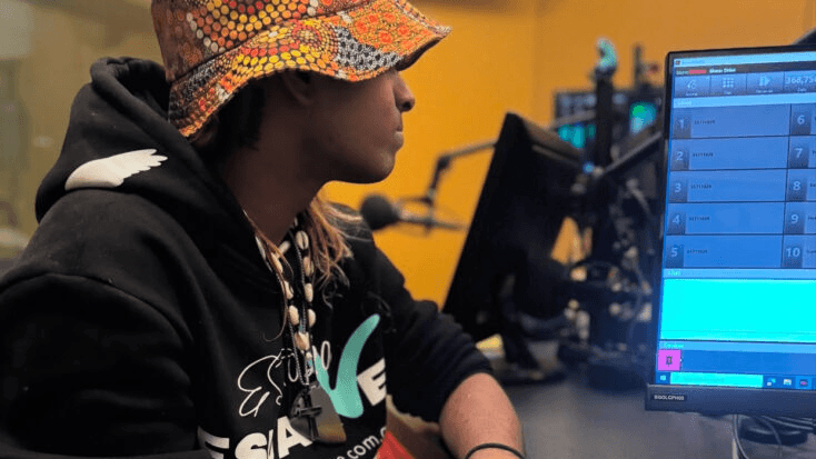 Person in a colorful bucket hat speaking into a microphone at a radio studio computer setup.