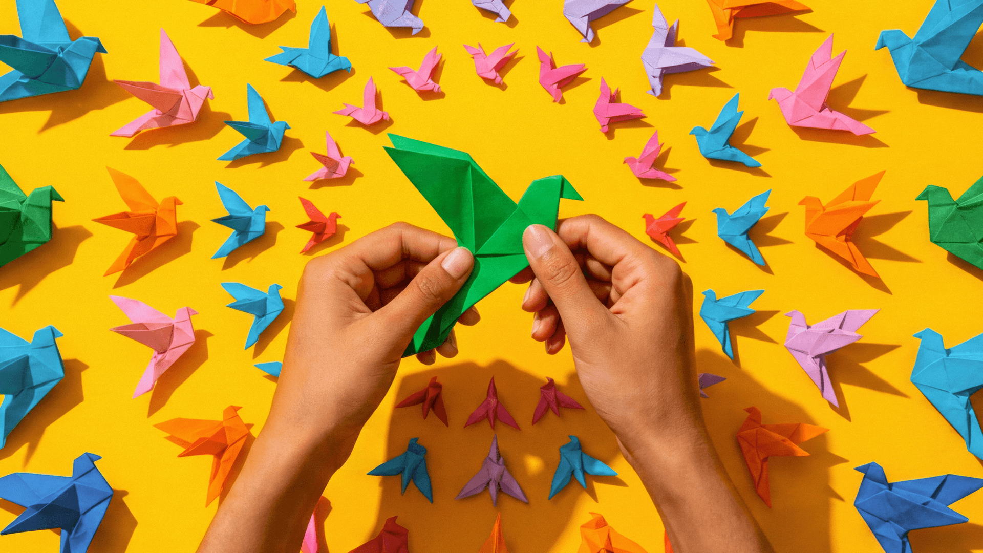 Hands folding a green origami bird amid colorful paper birds on a yellow background