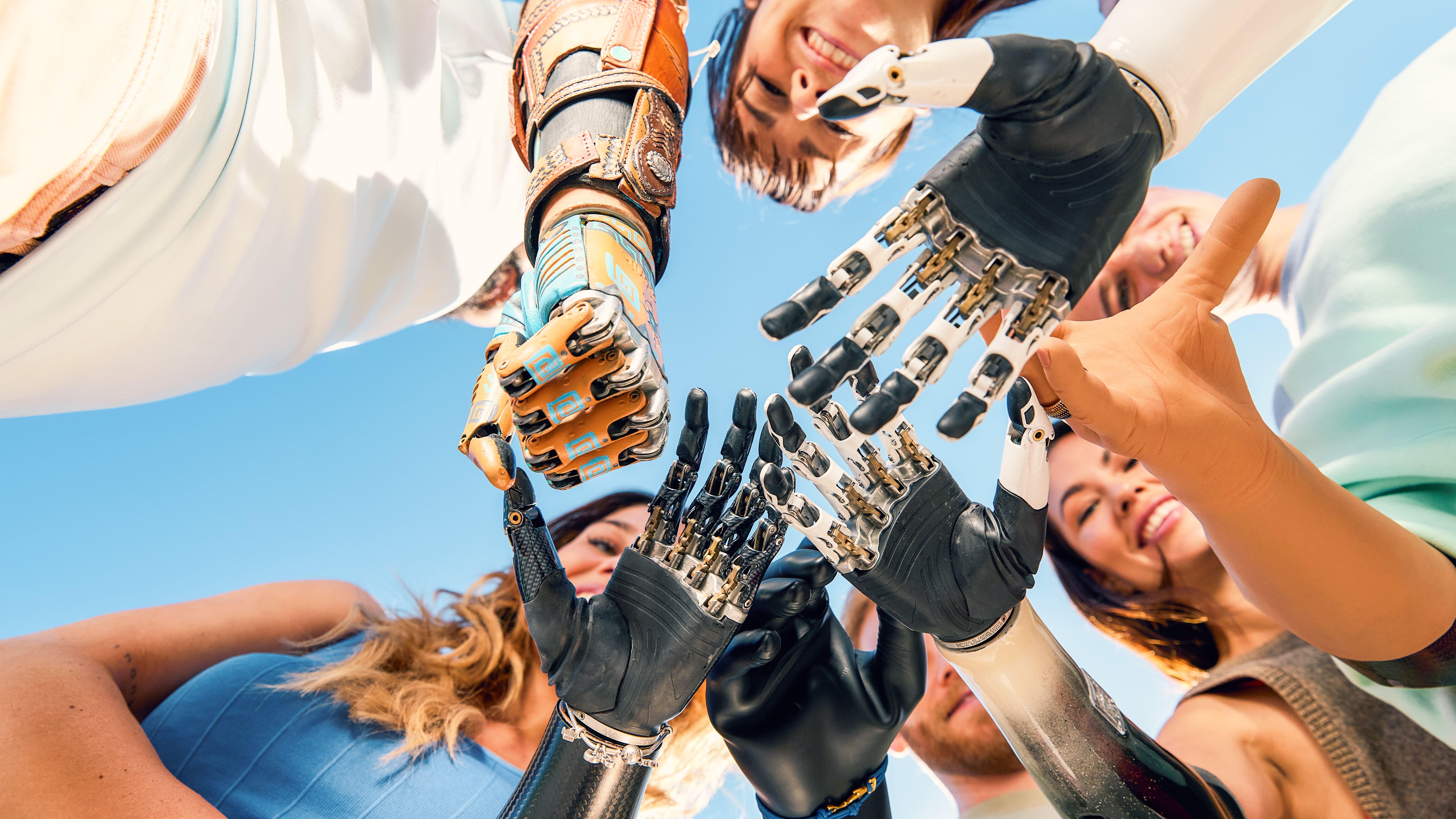 Close-up of mixed human and prosthetic hands stacked together in a circle against a blue sky.