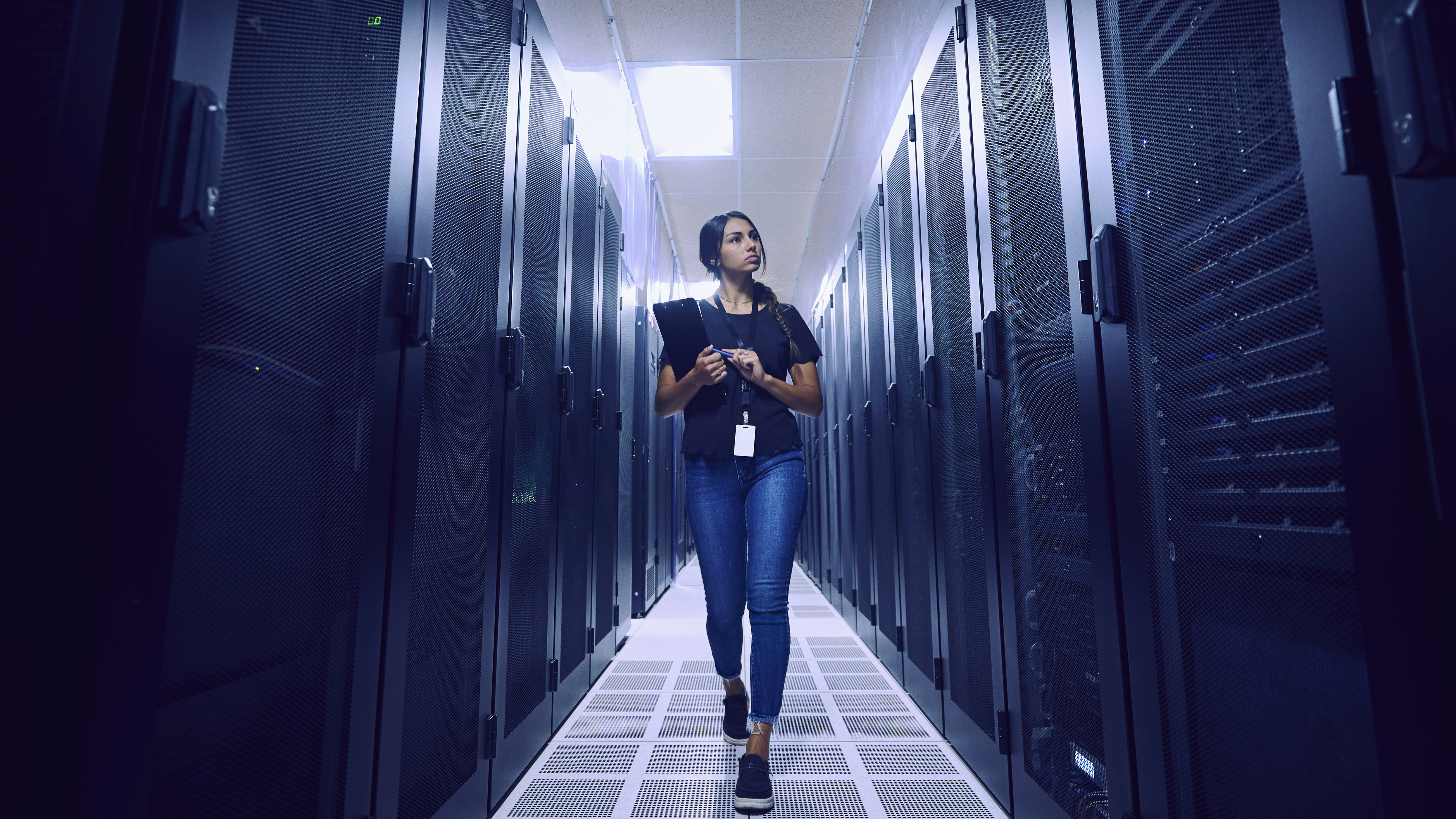 woman walking through a server room aisle holding a tablet.