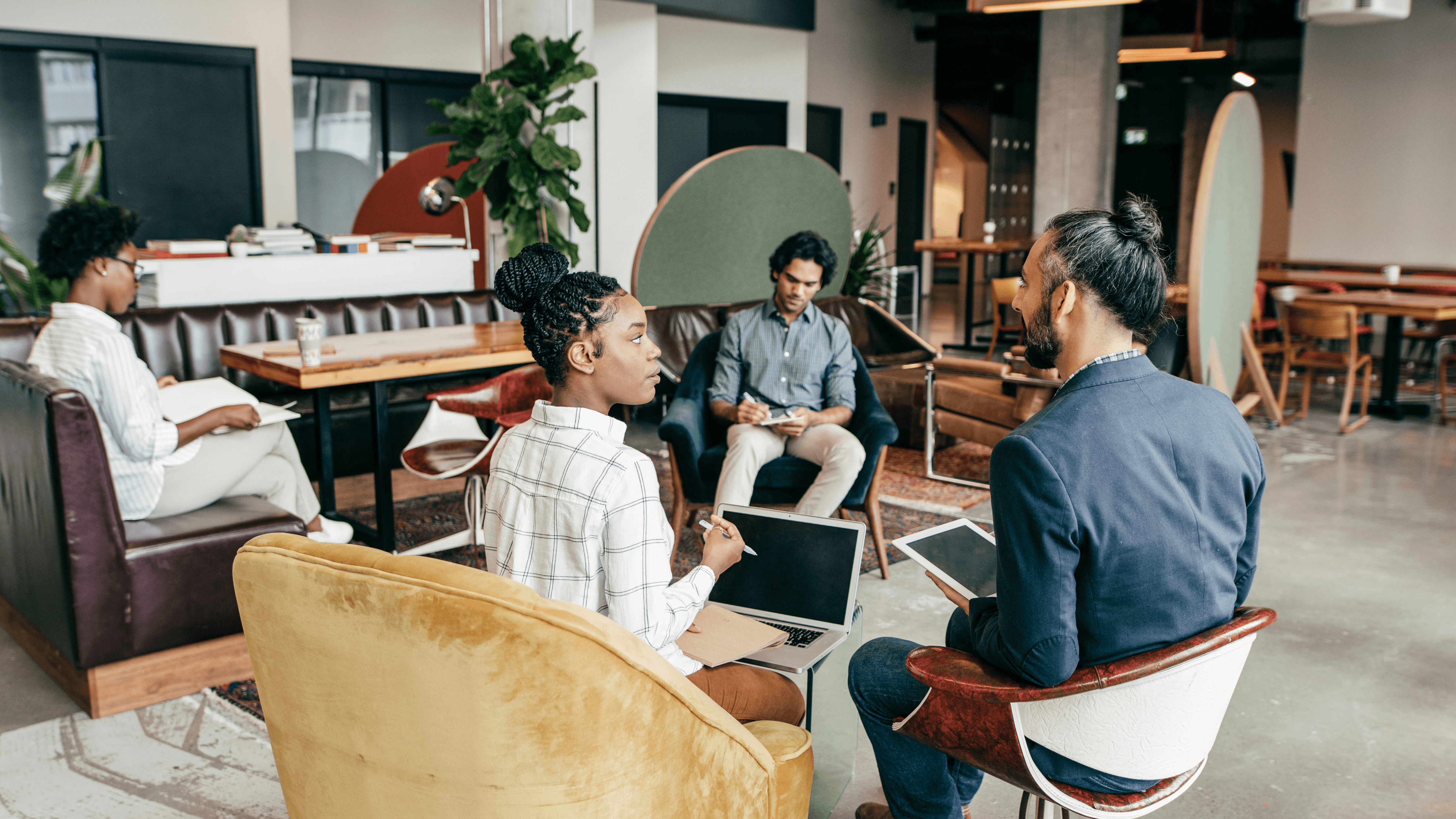 Small group seated in lounge chairs, discussing work with laptops and notebooks in a modern office space.