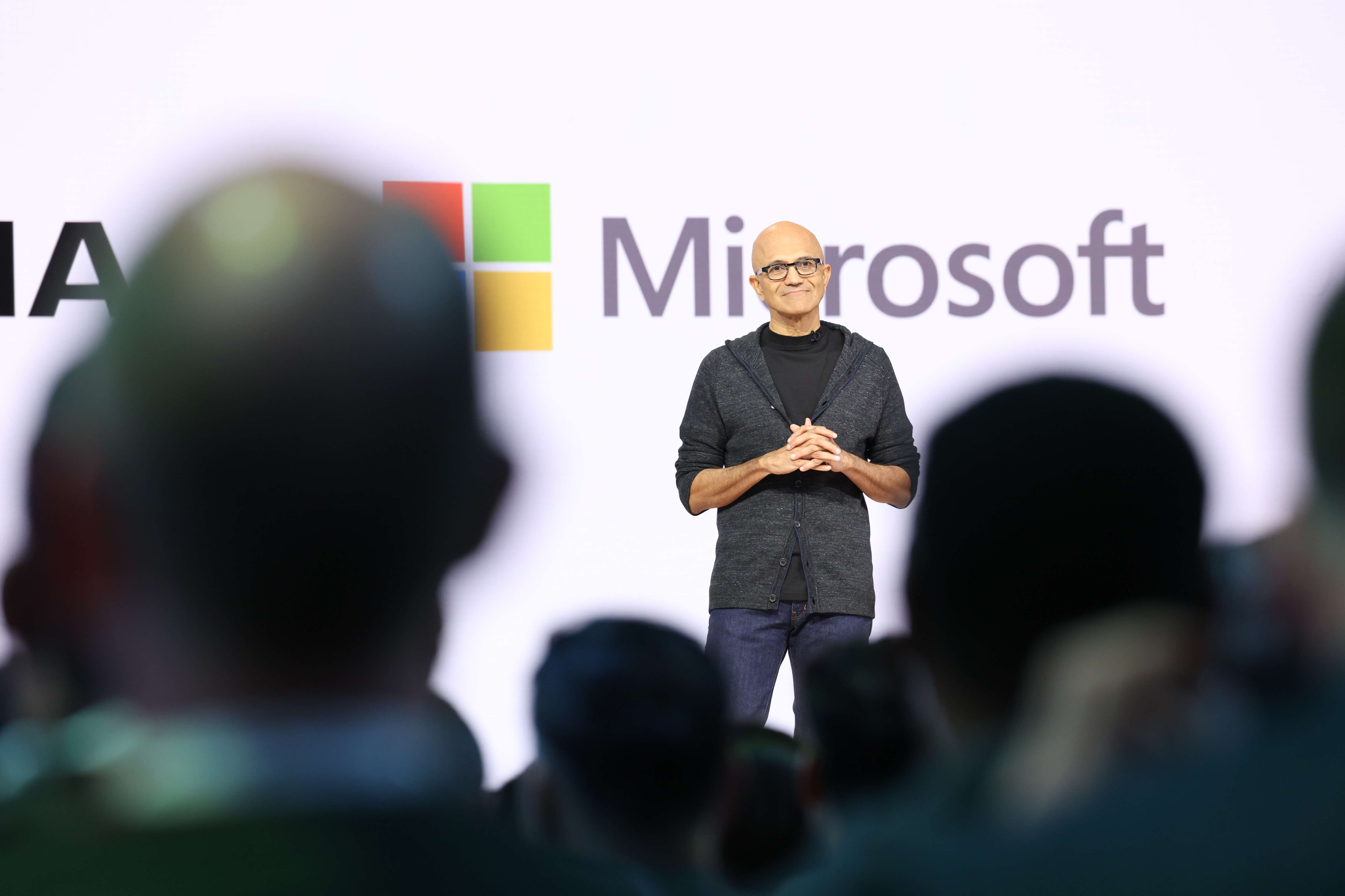 Satya Nadella speaking onstage with Microsoft logo behind, audience silhouettes in foreground.