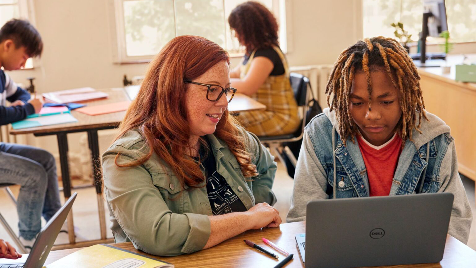 Teacher helping a student with a laptop in a classroom while others work in the background.
