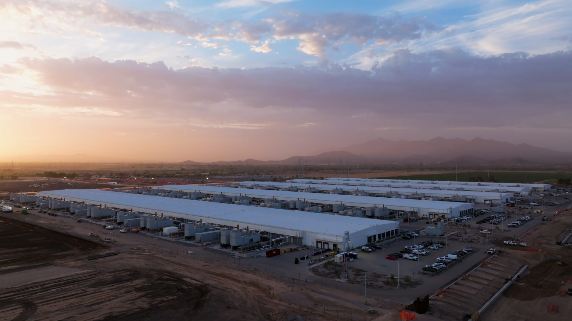Aerial view of a large industrial facility at sunrise, with rows of white-roofed buildings set against a desert landscape and distant mountains.