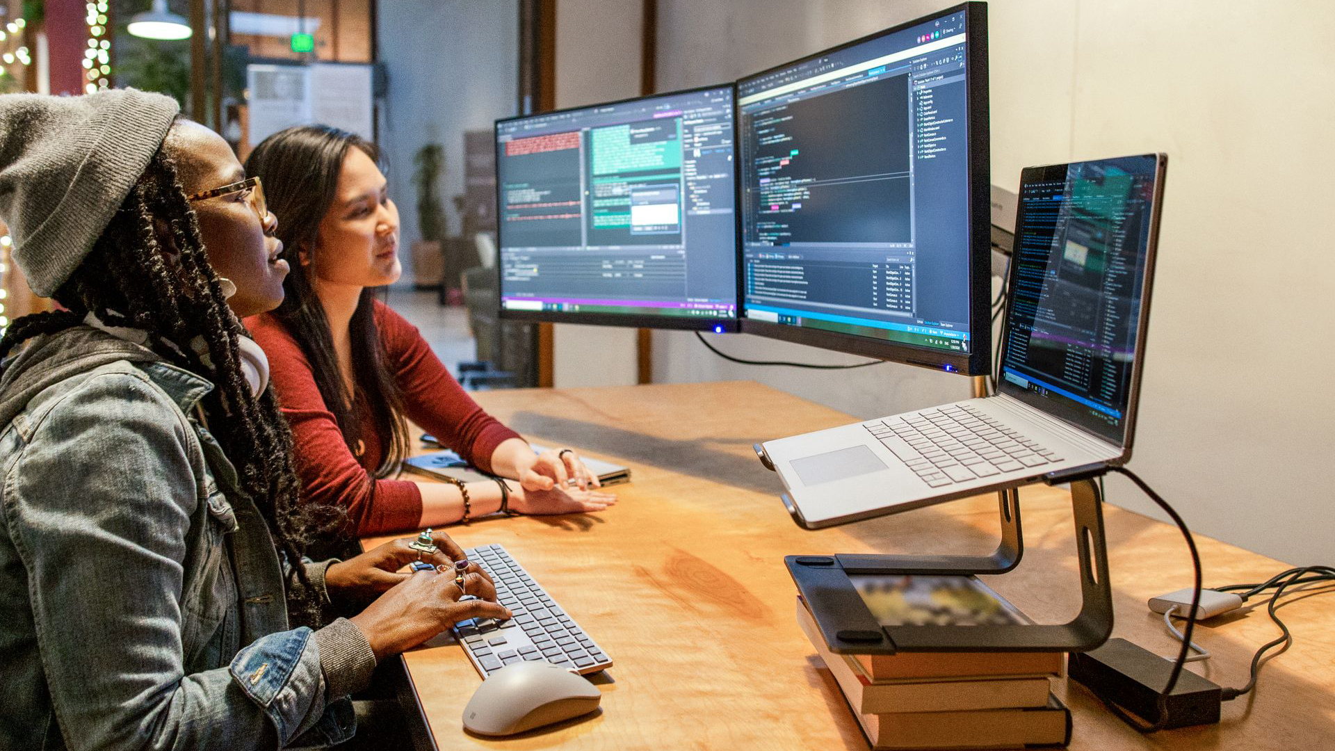 Two people working at a desk with dual monitors and a laptop, reviewing and writing code.