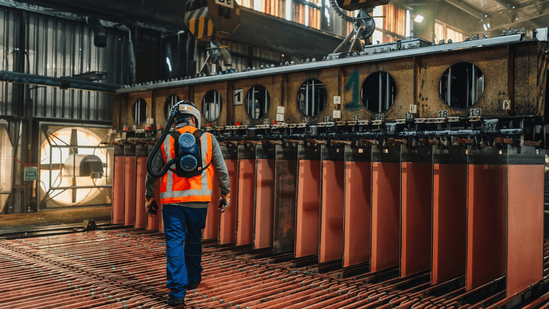Worker in safety gear with a respirator walking past large industrial processing equipment in a factory.