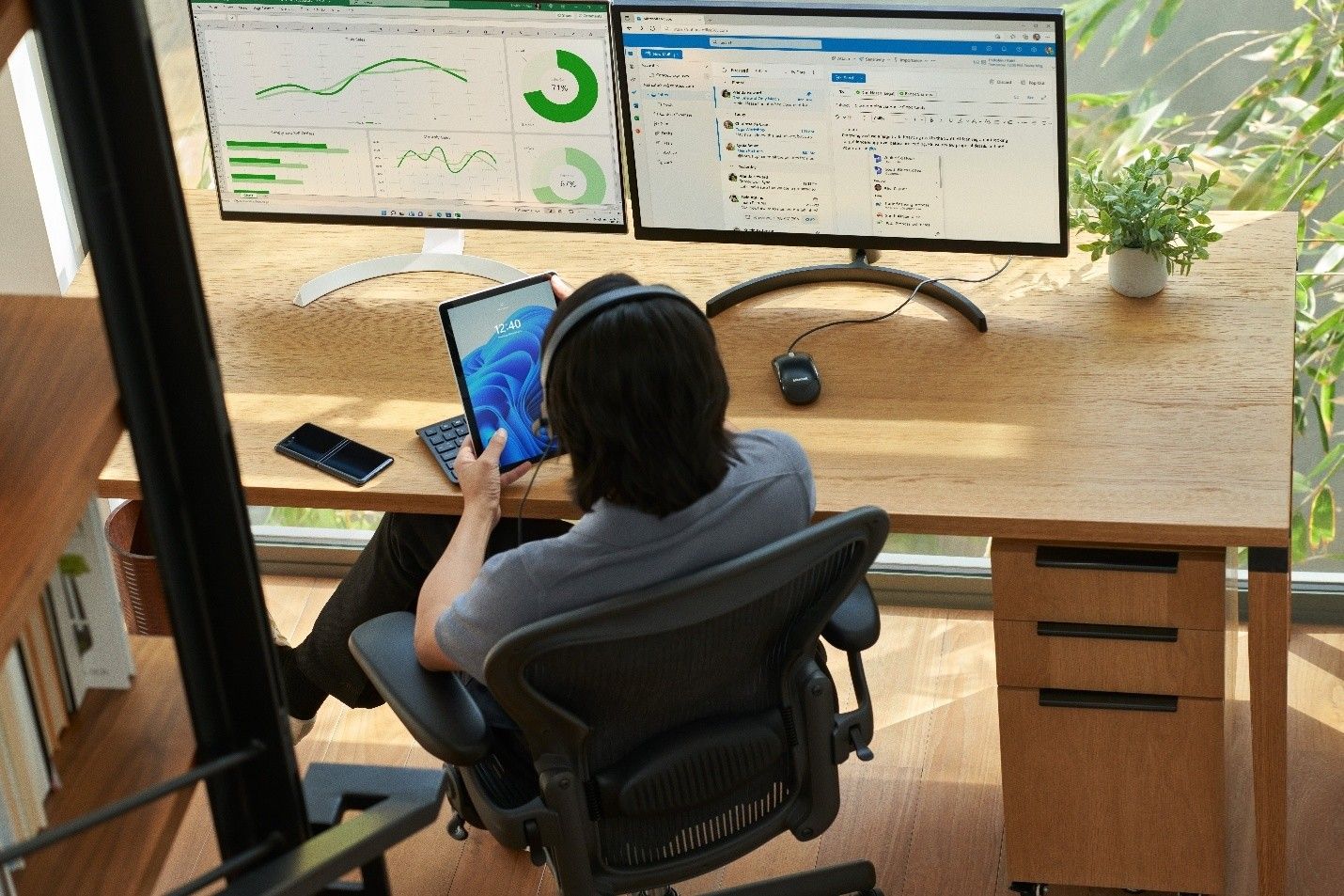 Person at desk using a tablet with dual monitors showing data dashboards and email interface.