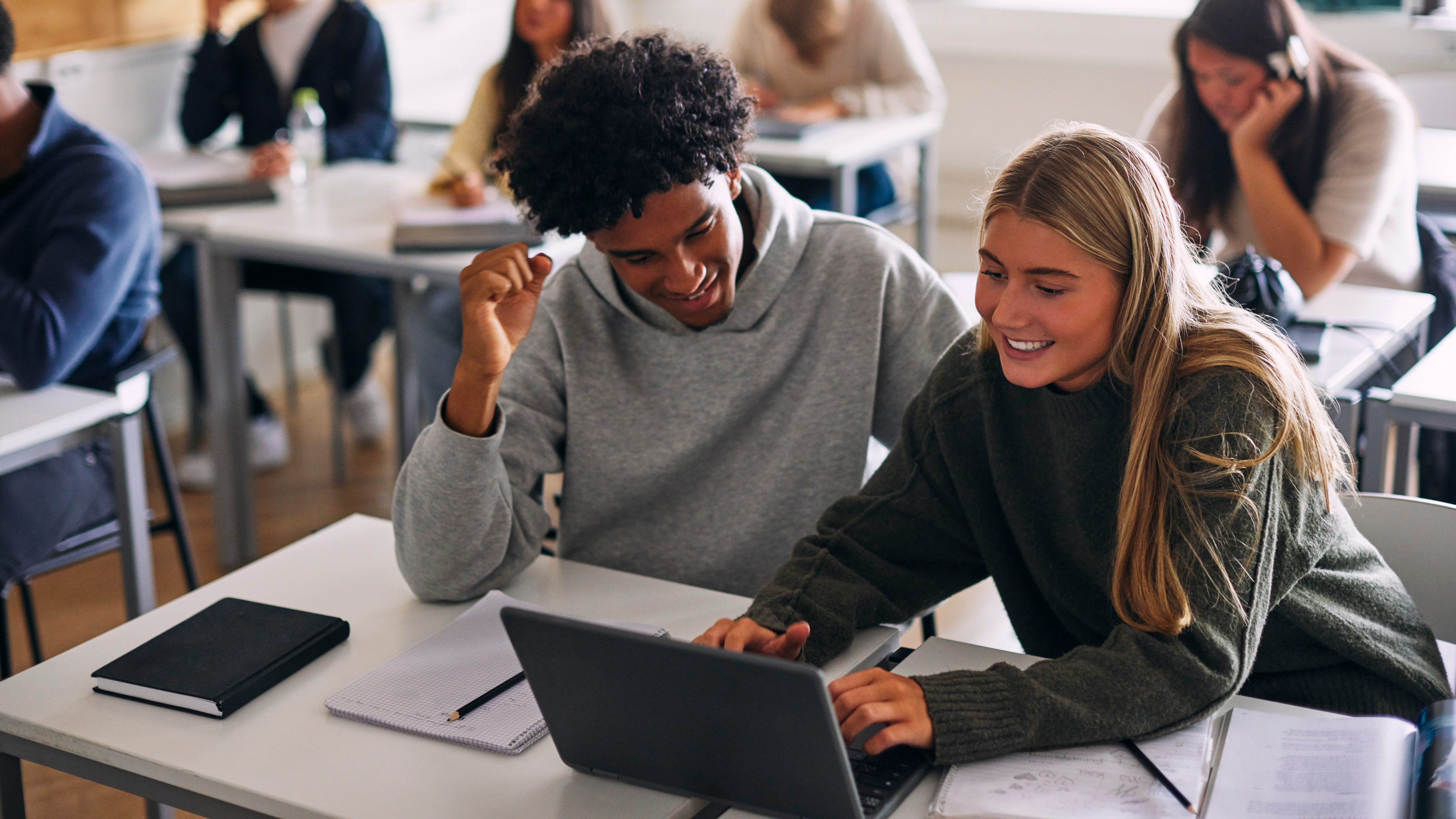 Two students sitting at desks in a classroom, working together on a laptop.