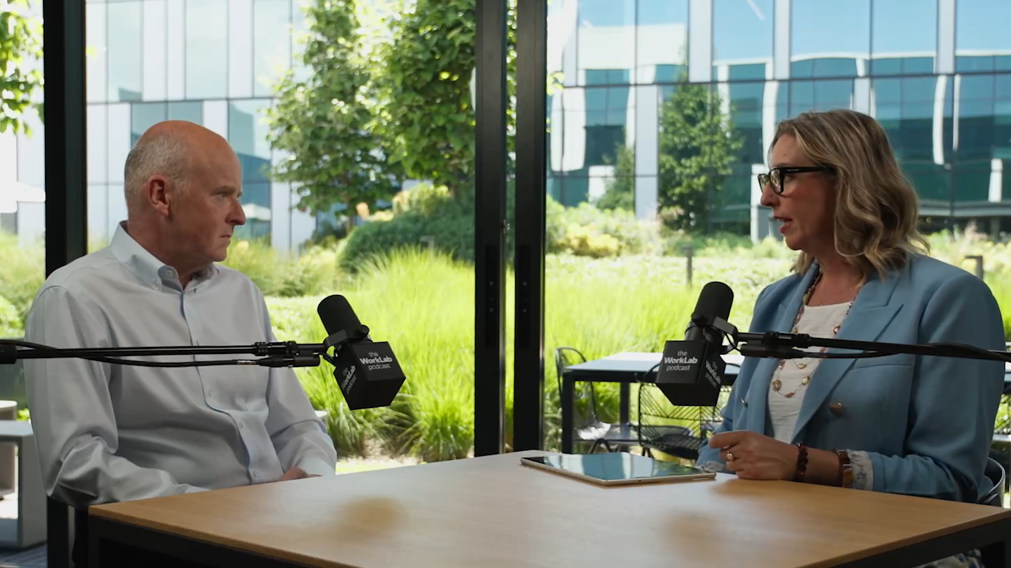 Two people seated at a table, speaking into microphones in a podcast-style interview setting.
