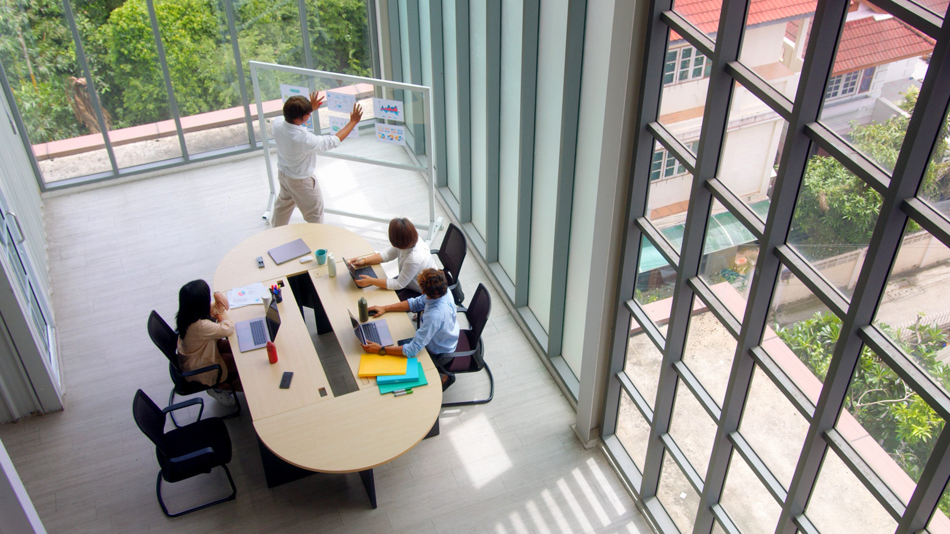 Team members collaborate in a glass-walled conference room while a colleague presents at a whiteboard.