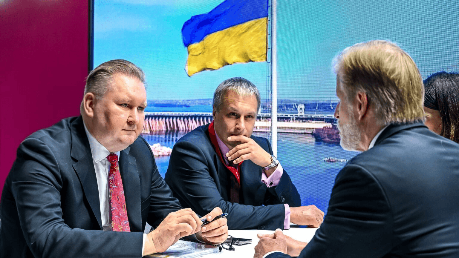 People seated at a table in discussion, with a large Ukrainian flag displayed on a screen in the background.