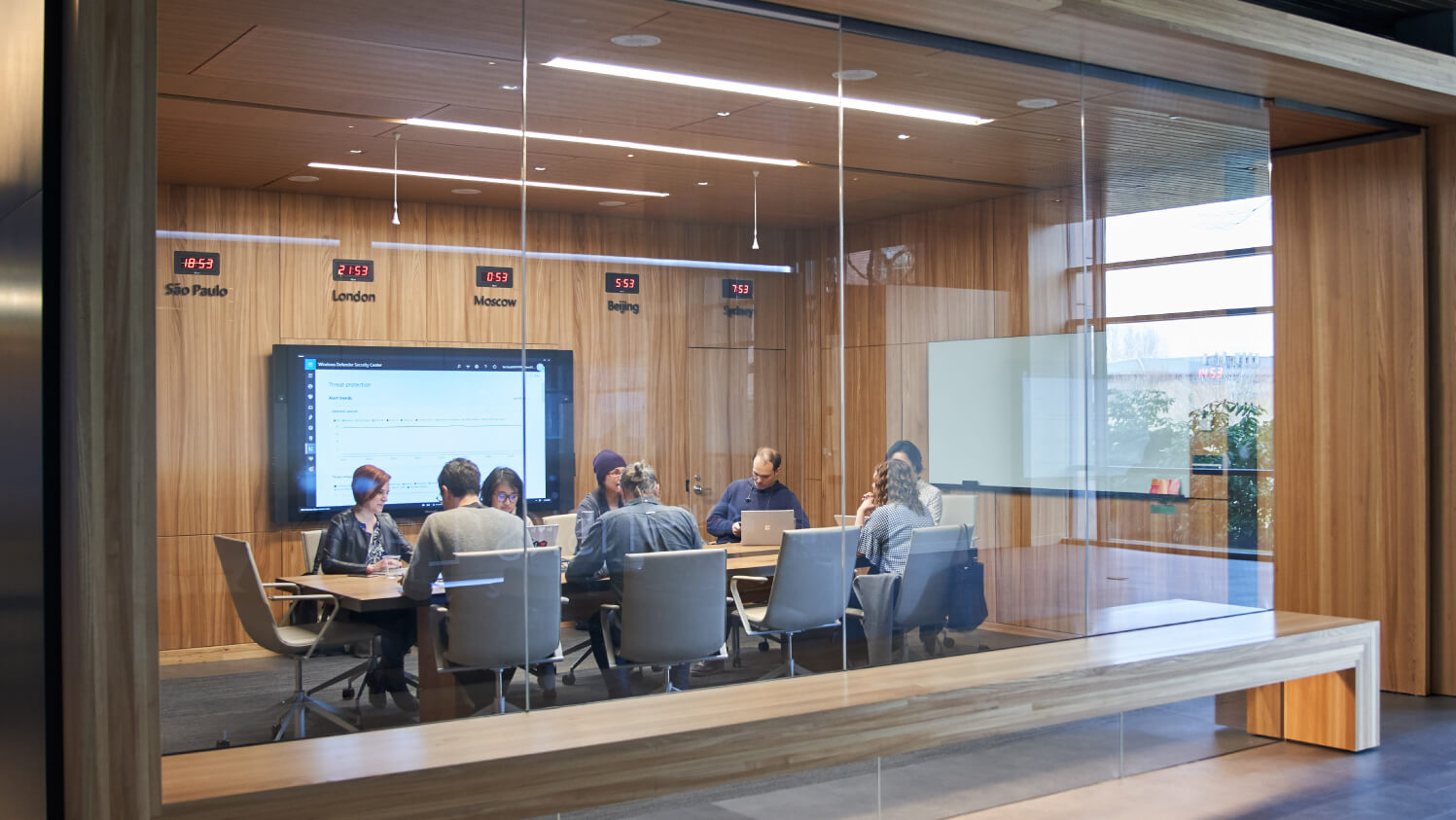 People sit around a conference table in a glass-walled meeting room, working on laptops and discussing information displayed on a large screen, with world clocks mounted on the wood-paneled wall behind them.