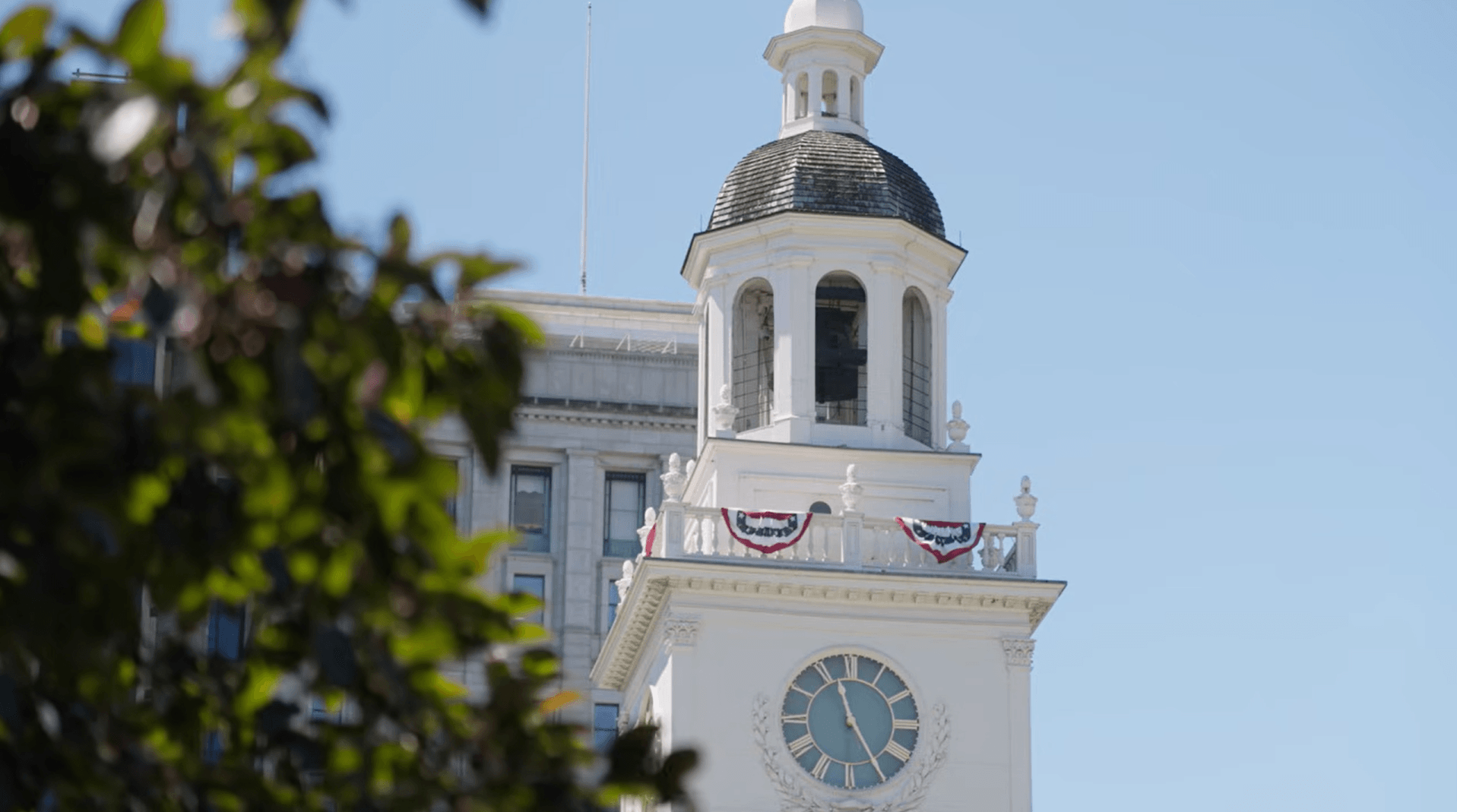 Historic clock tower decorated with patriotic bunting, framed by trees against a clear blue sky.