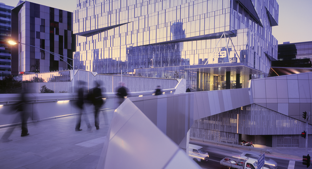 Modern glass office building with illuminated walkway and blurred pedestrians at dusk.