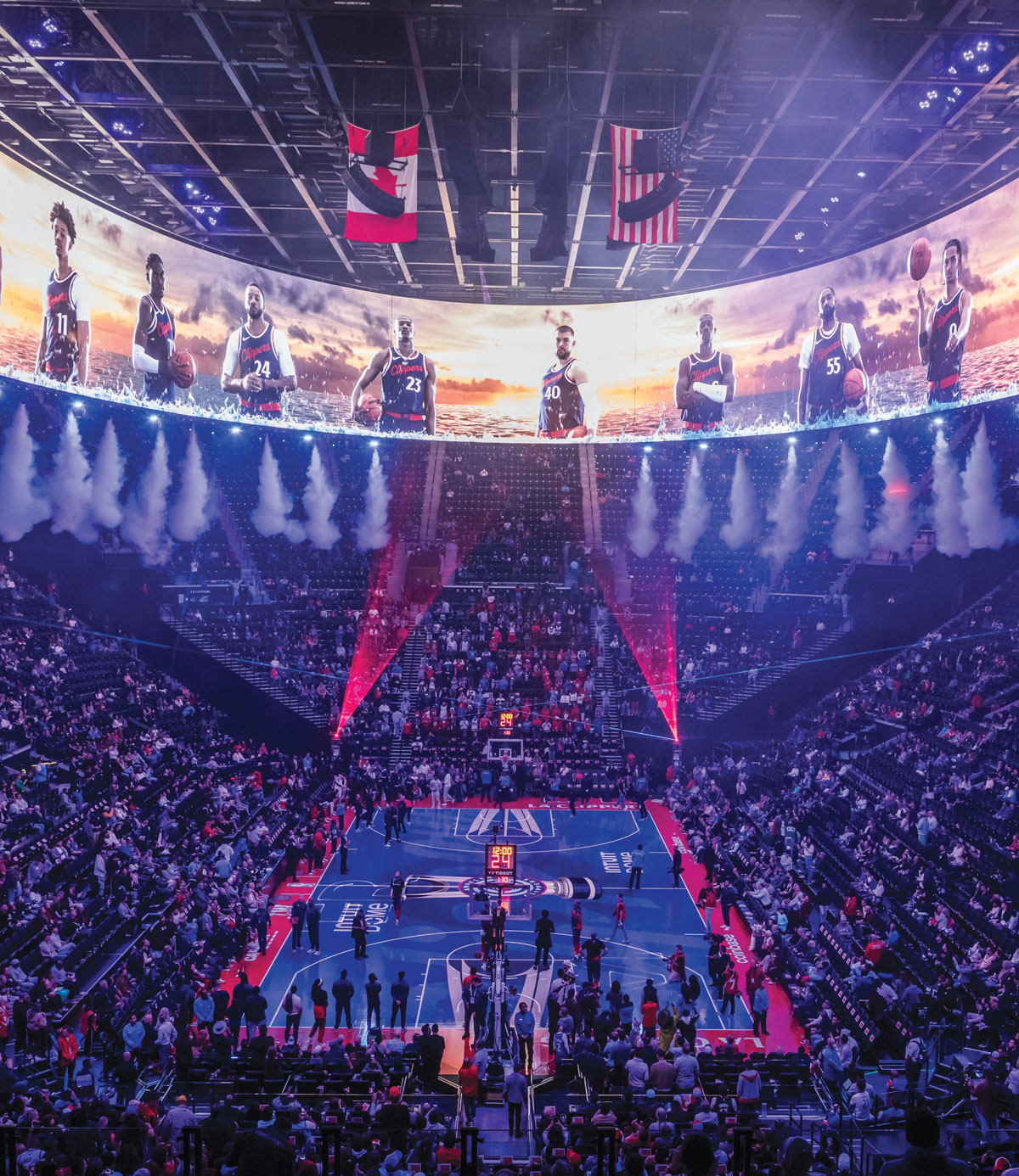 A packed basketball arena with dramatic lighting and smoke effects, featuring player images on a large curved screen above the court as teams prepare for the game. The crowd fills the stands, and flags hang from the ceiling.