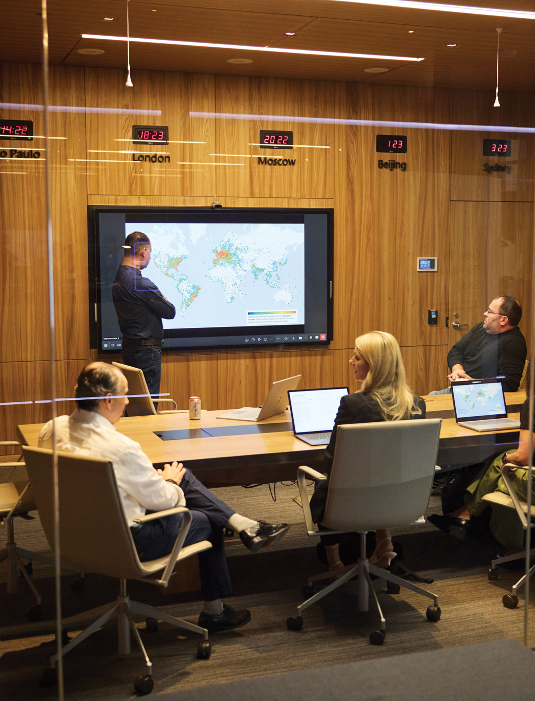 A man presents a world map on a large screen to four seated colleagues in a modern conference room. Laptops are open on the table, and world clocks are mounted on a wood-paneled wall.