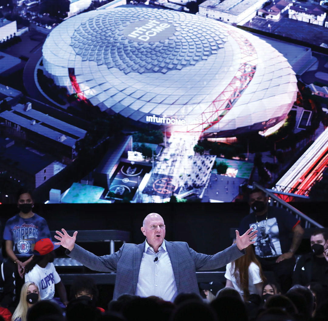 A man stands with arms outstretched in front of a seated audience, with a large screen behind him displaying an aerial rendering of the Intuit Dome sports arena.