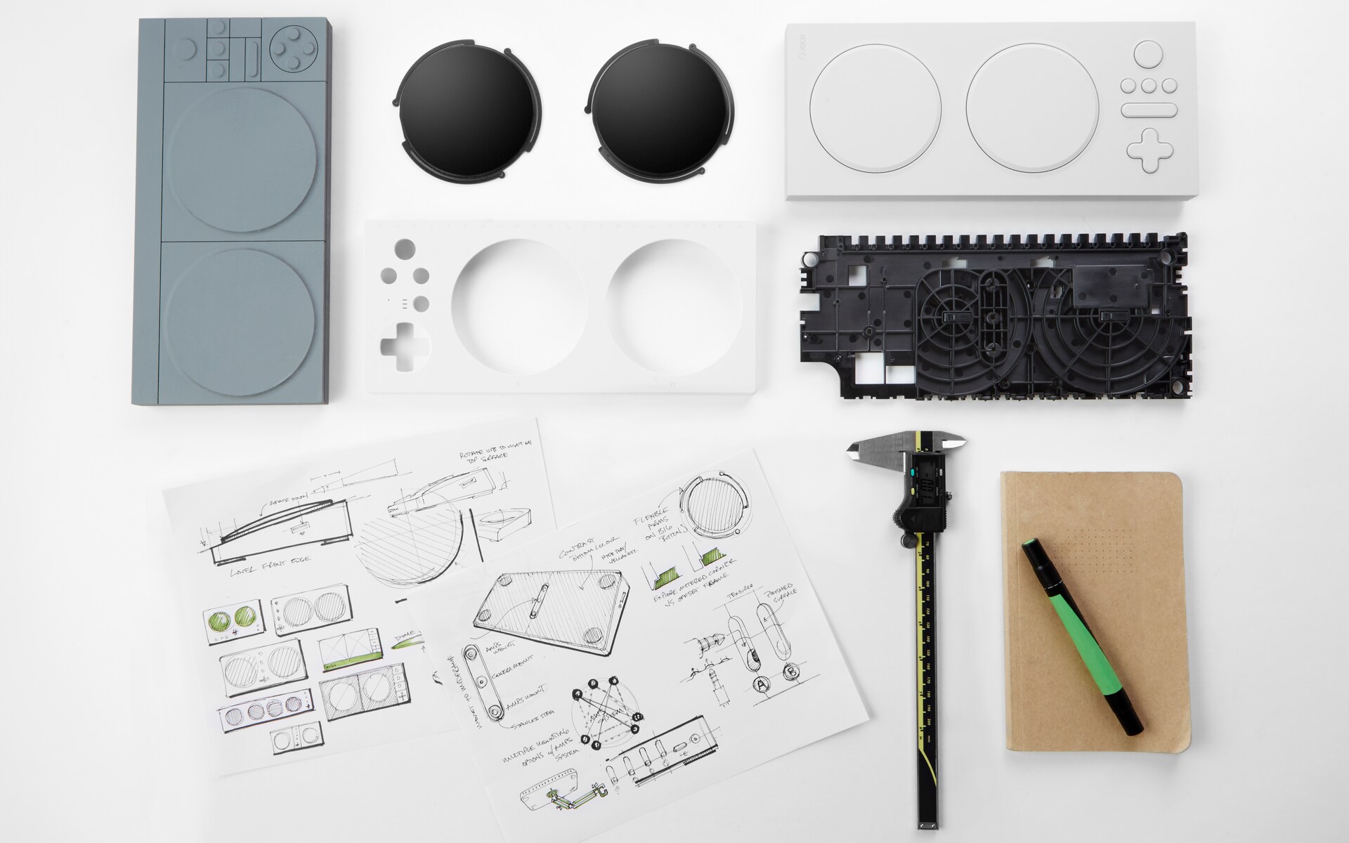 A workspace with sketches, a caliper, pen, notebook, and various components of an adaptive game controller, including buttons, circuit board, and casing parts, arranged neatly on a white surface.