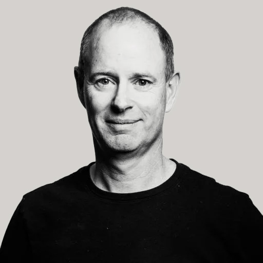 A middle-aged man with short hair and a slight smile, wearing a black crew-neck shirt, poses against a plain light gray background.