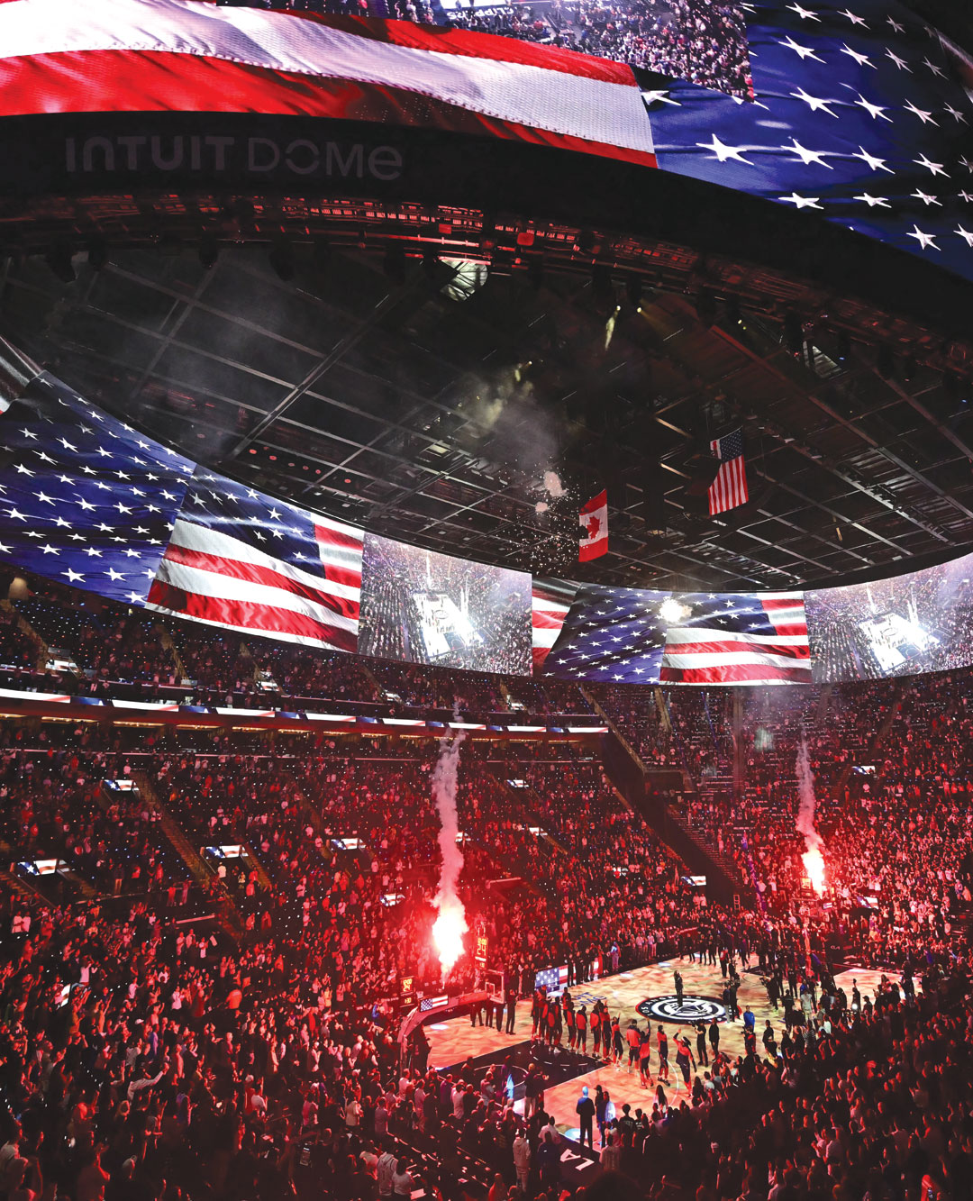 A packed basketball arena with a large digital display showing the American flag overhead. The court is lit, smoke effects rise, and players stand lined up for a pre-game ceremony. The crowd fills the stands.