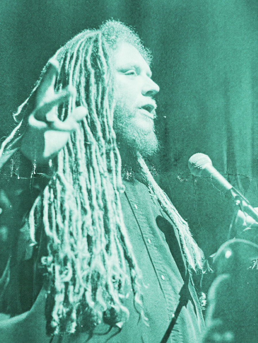 A person with long, light-colored dreadlocks and a beard speaks passionately into a microphone, gesturing with one hand. The image has a teal color filter and a textured, vintage effect.