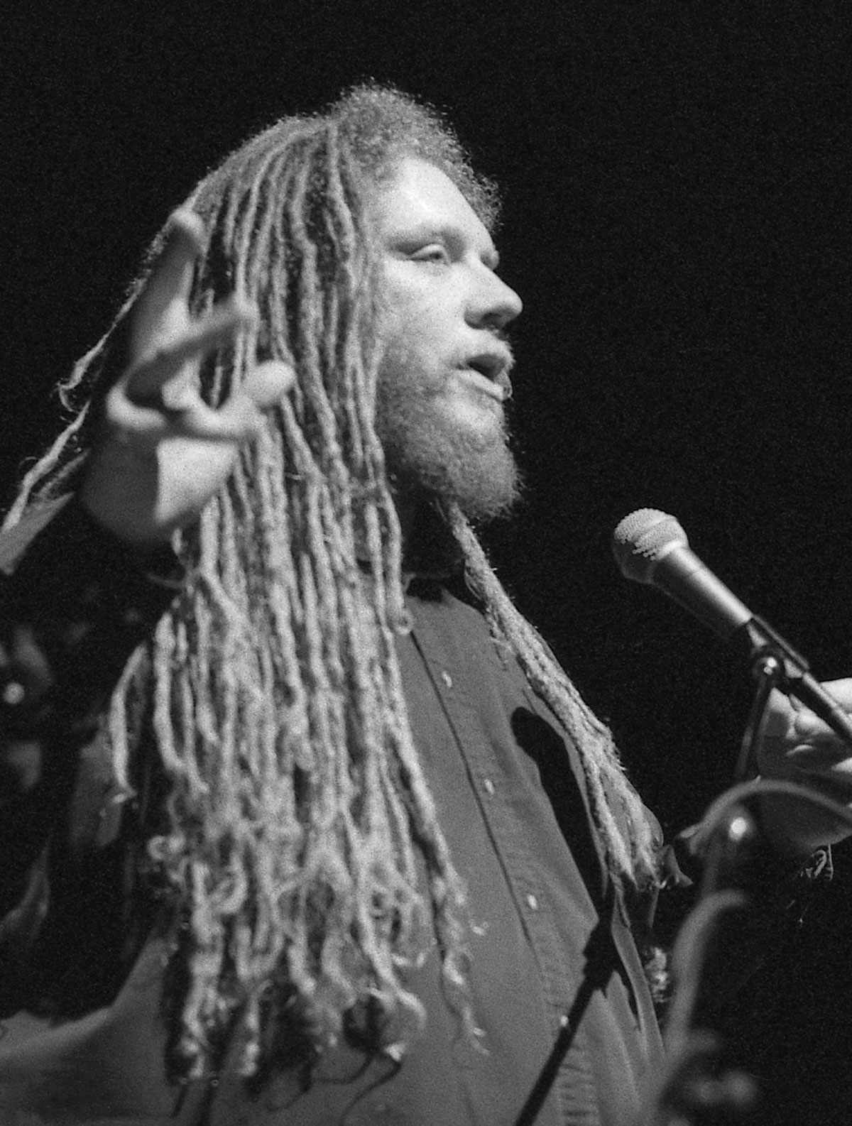 A person with long dreadlocks and a beard speaks passionately into a microphone, gesturing with one hand in front of a dark background.