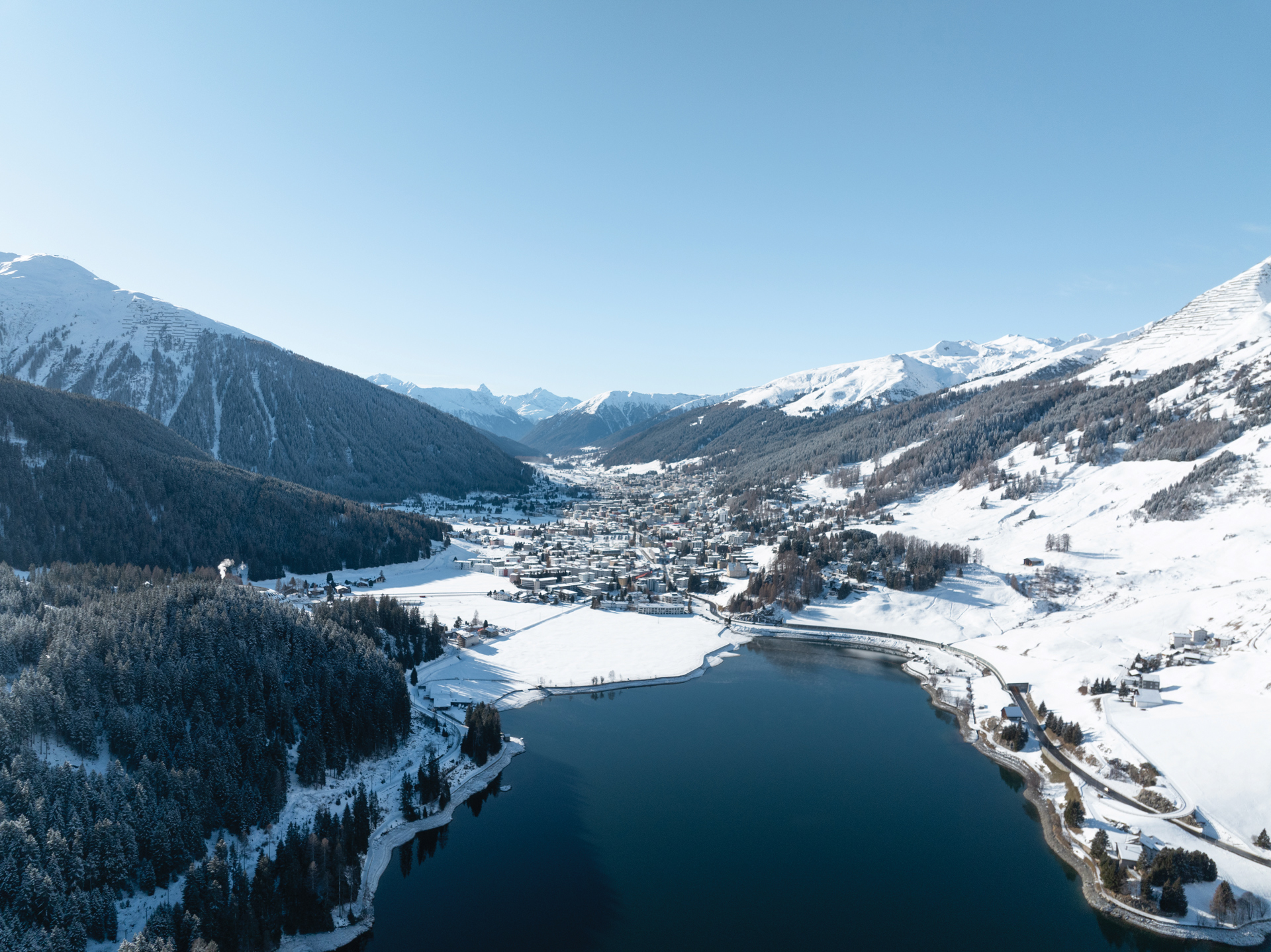 A snowy mountain town sits beside a lake, surrounded by pine forests and snow-covered slopes under a clear blue sky.