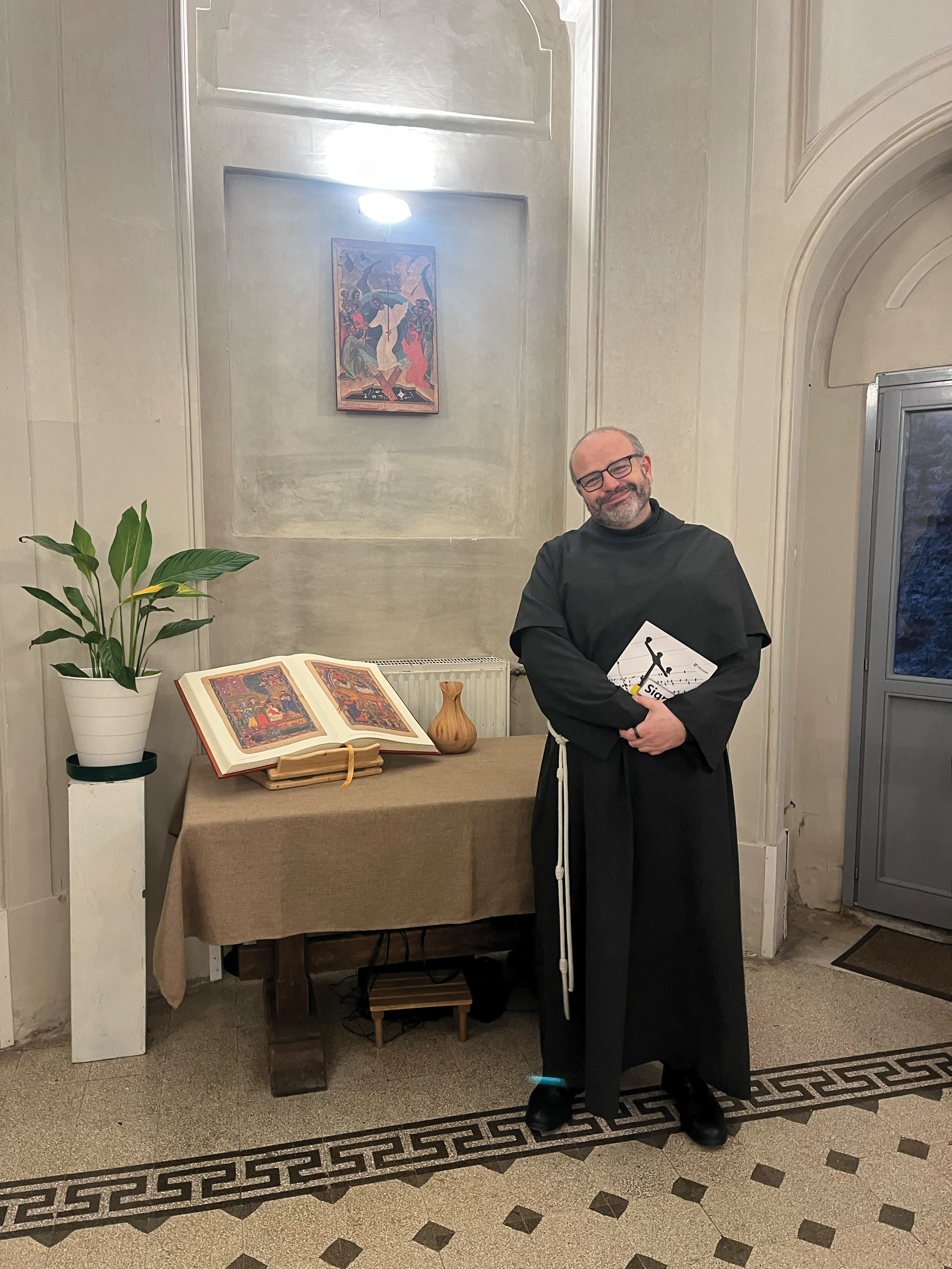 A smiling man in a black religious habit stands beside a table holding an open illustrated book, a vase, and a potted plant in a well-lit room with a decorative tile floor and wall art.