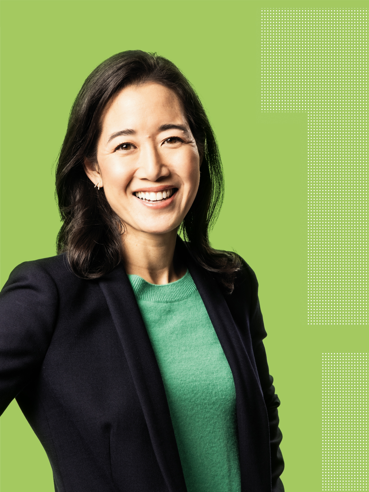 A woman with dark hair wearing a green top and a dark blazer smiles against a light green background with a dotted design on the right side.