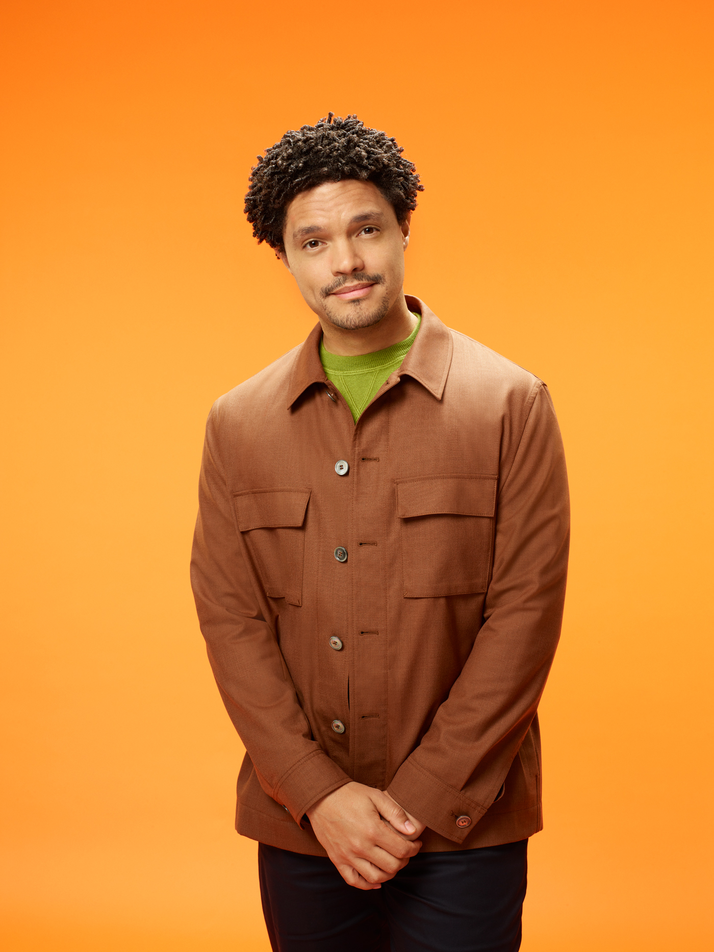 A man with curly hair stands against an orange background, wearing a brown button-up jacket over a green shirt. He is looking at the camera with a slight smile, hands loosely clasped in front of him.