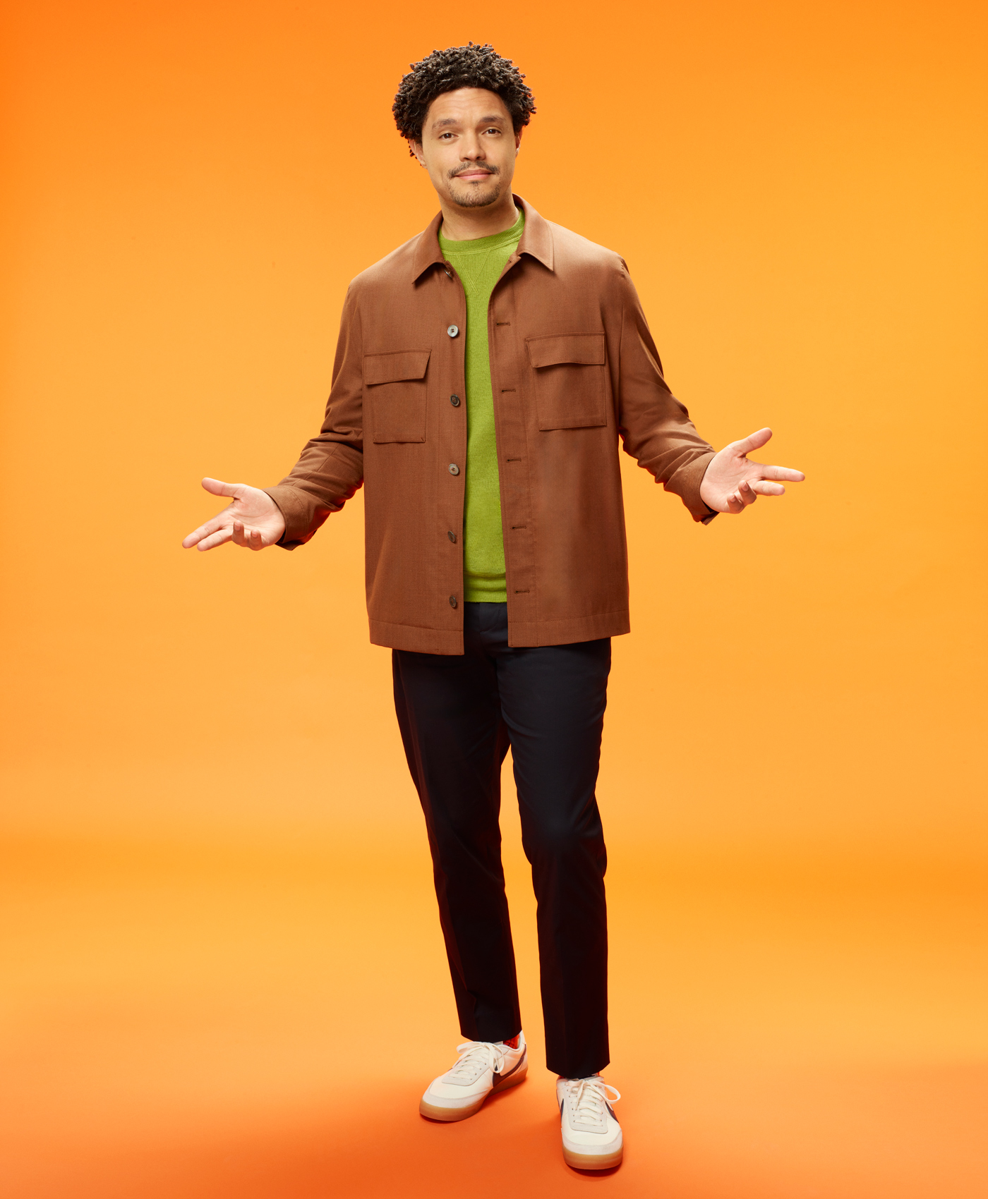 A man with short curly hair stands against an orange background, wearing a brown jacket over a green sweater, dark pants, and white sneakers. He is smiling and gesturing with his hands open to the sides.