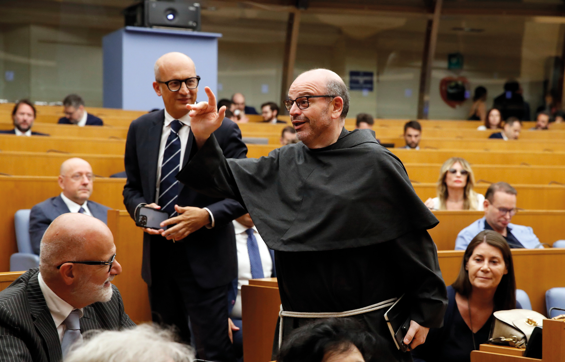 A man wearing a black robe and glasses gestures while standing in a crowded auditorium, surrounded by seated people in formal attire. The setting appears to be an official or formal meeting.