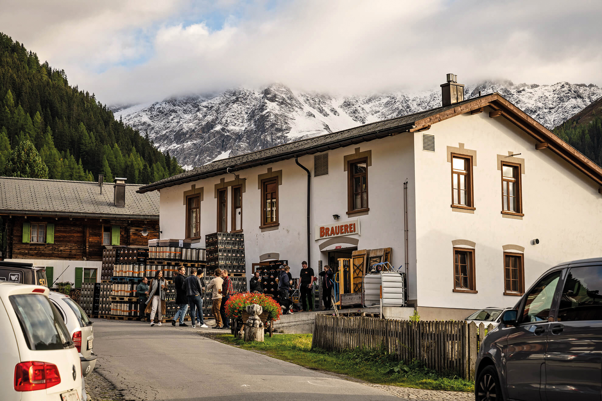 A group of people gathers outside a brewery in a quaint alpine village, with snow-capped mountains and evergreen trees in the background under a cloudy sky. Cars are parked along the narrow road.