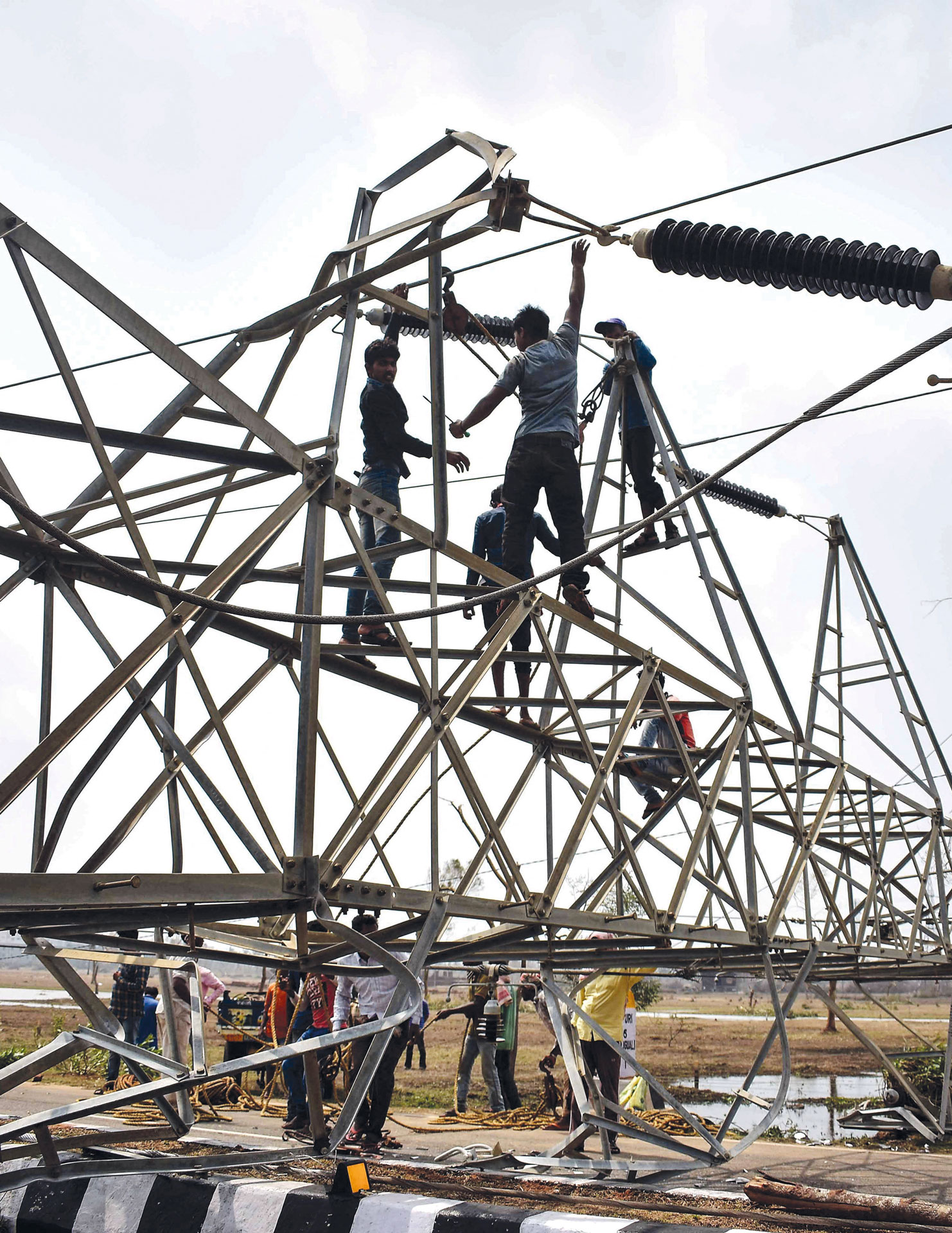 Workers climb and repair a tilted metal electricity tower, with others gathered below on the ground. The scene is outdoors in a rural area, under a cloudy sky.