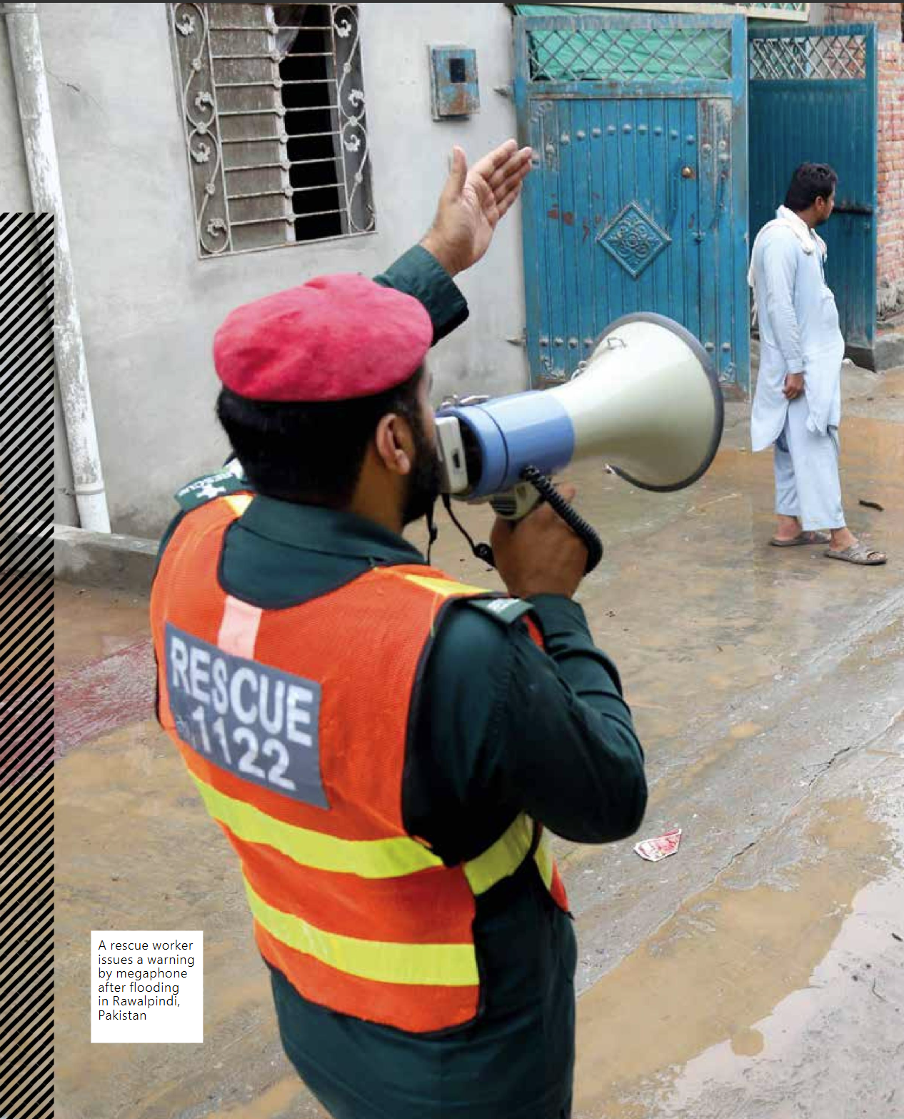 A rescue worker in a red beret and orange vest uses a megaphone to address residents on a flooded street in Raiwind, Pakistan. A man in traditional clothing walks past a blue gate in the background.