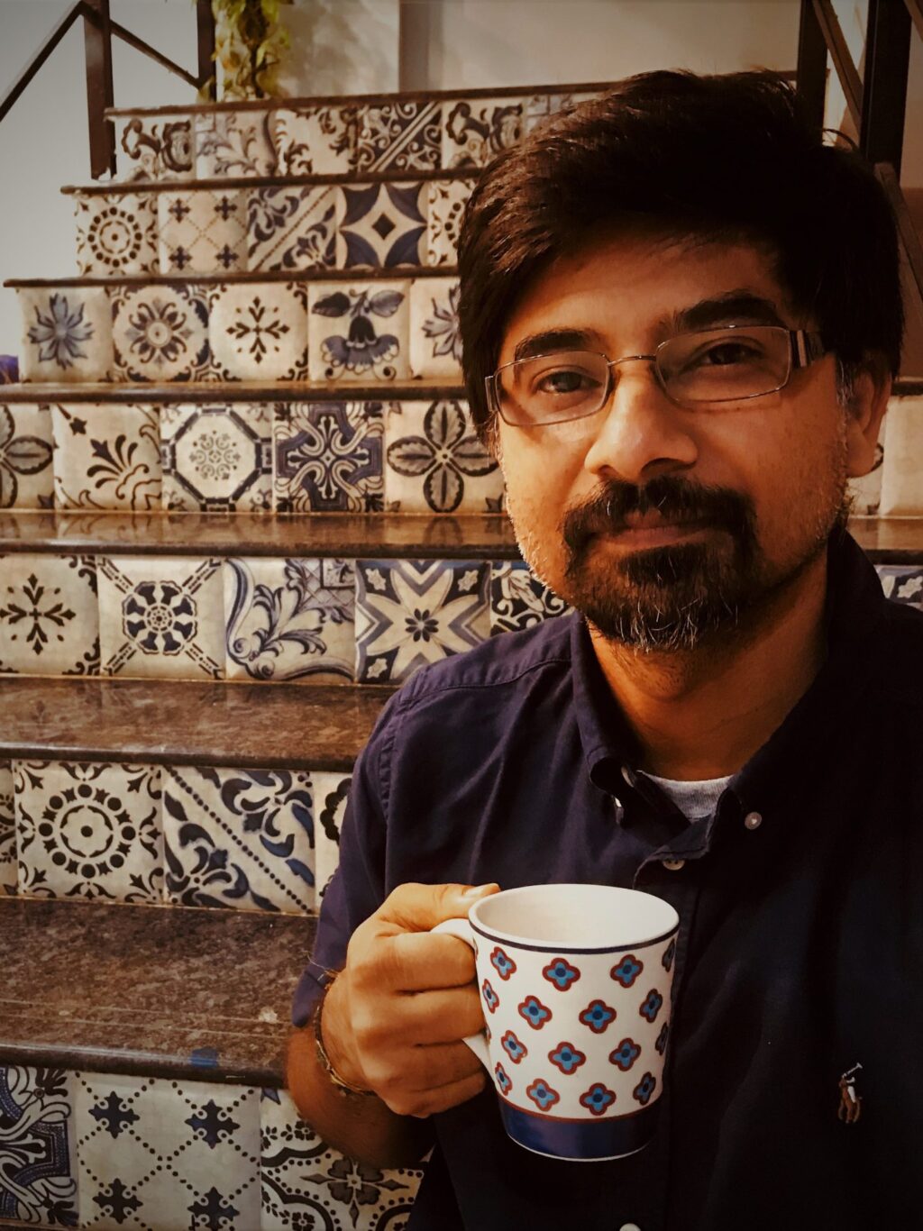 a man with black hair, wearing glasses and a beard, holding a coffee cup