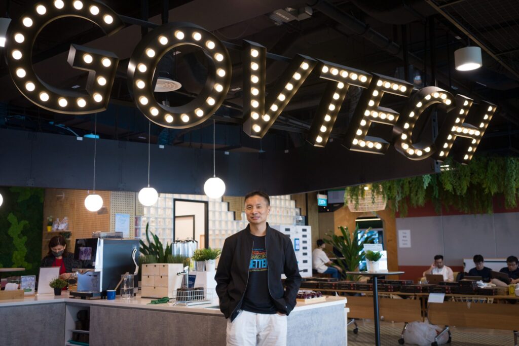 A man casually posing against a desk under a lit signage that reads GOVTECH