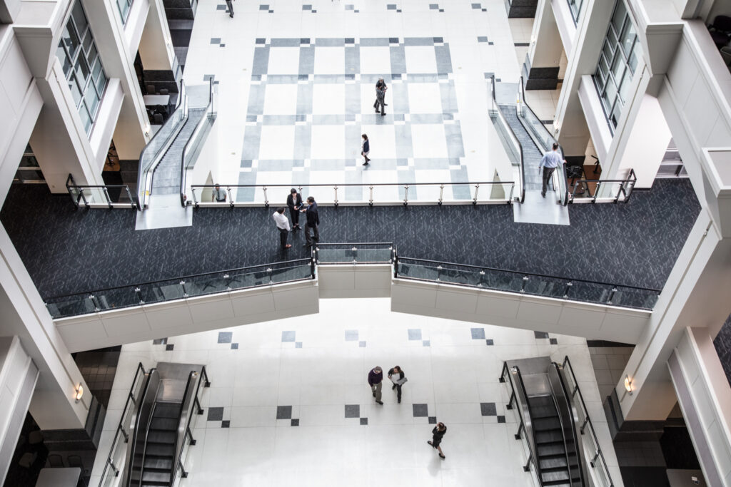 A high-angle view of a modern, multi-story atrium with people walking and conversing. The atrium features large windows, escalators, and a checkered floor pattern. The image captures the spaciousness and architectural design of the building.